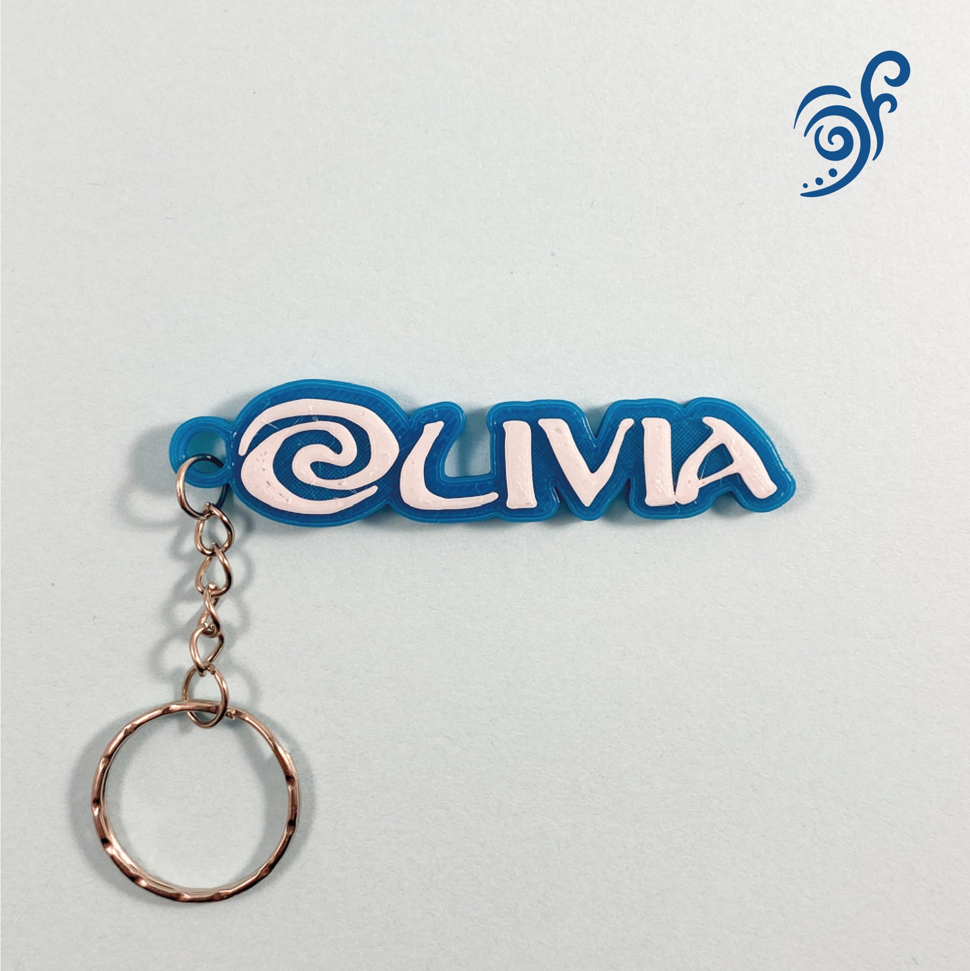 Porte-clés personnalisé avec le prénom 'Olivia' en relief blanc sur une base bleue, avec un motif de spirale océanique décorative à la place du O. Inspiré de l'Aventure Océanique de Vaiana, sur fond gris clair. Un motif de vagues bleues est visible dans le coin supérieur droit.
