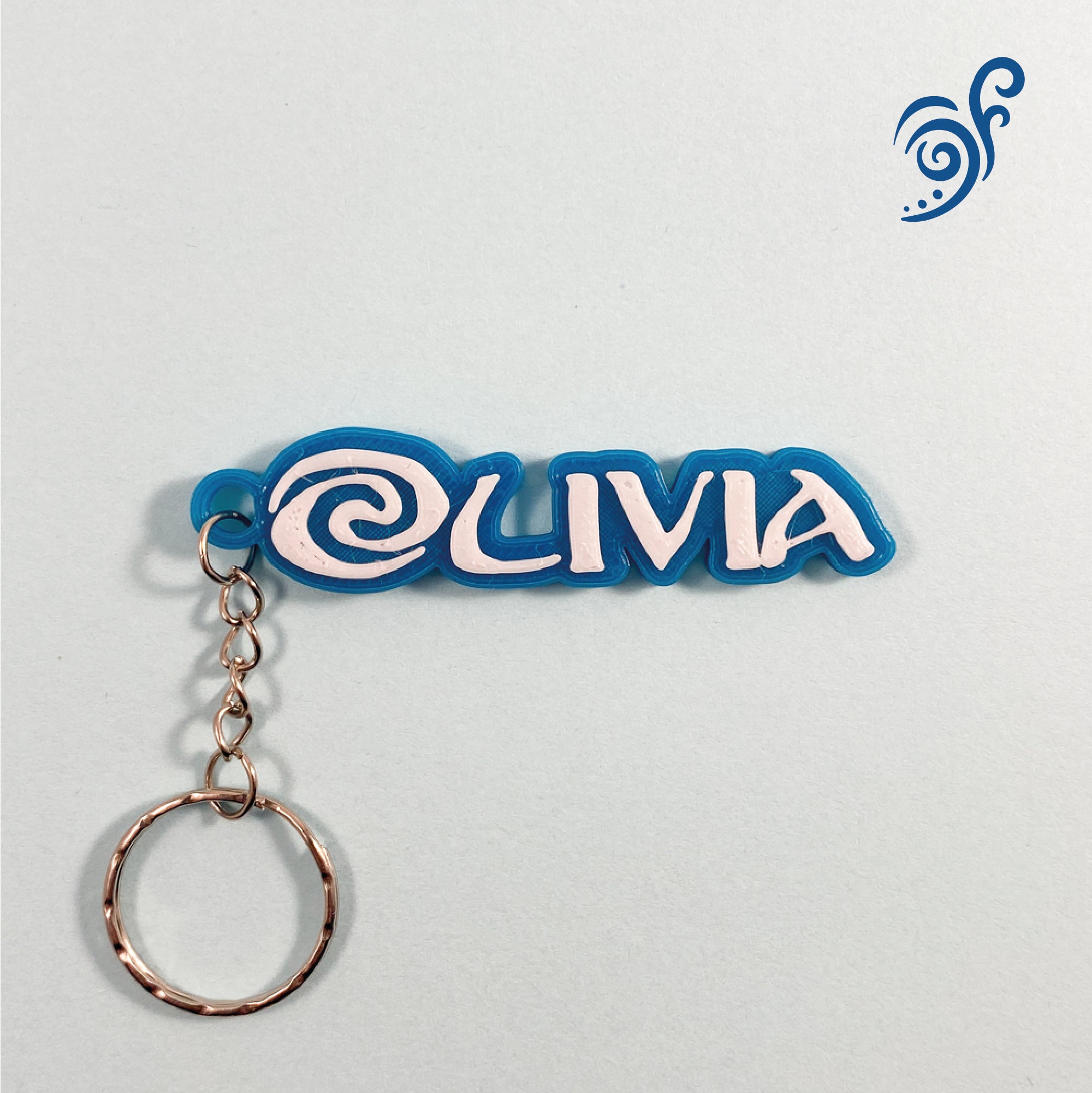 Porte-clés personnalisé avec le prénom 'Olivia' en relief blanc sur une base bleue, avec un motif de spirale océanique décorative à la place du O. Inspiré de l'Aventure Océanique de Vaiana, sur fond gris clair. Un motif de vagues bleues est visible dans le coin supérieur droit.