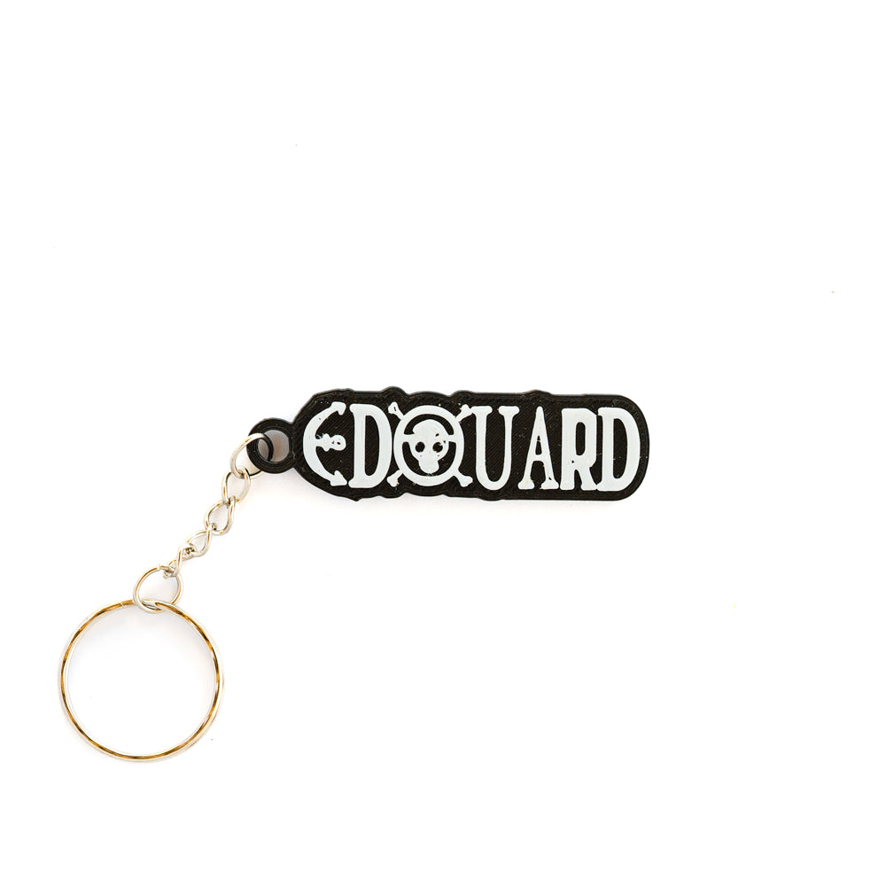 Porte-clés personnalisable noir et blanc sur fond uni, affichant le prénom 'EDOUARD' en police stylisée d'inspiration One Piece (Luffy), avec un petit emblème Jolly Roger (crâne) intégré entre les lettres D et O. L'attache est en métal argenté.