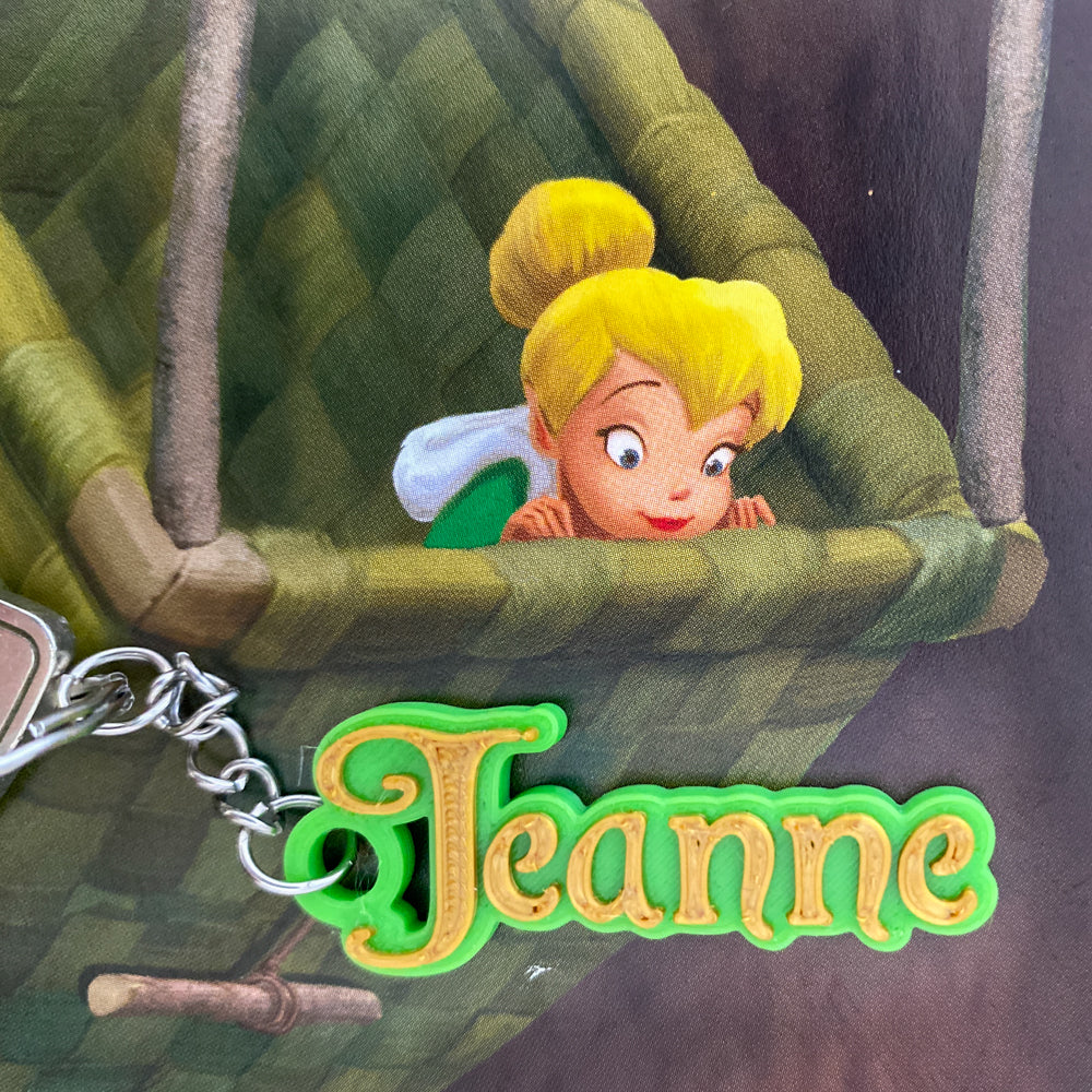 Porte-clés personnalisé au prénom Jeanne, avec le prénom en relief couleur or brillant sur un support vert vif. L'objet est présenté sur un fond illustré d'un panier en osier et de la Fée Clochette.