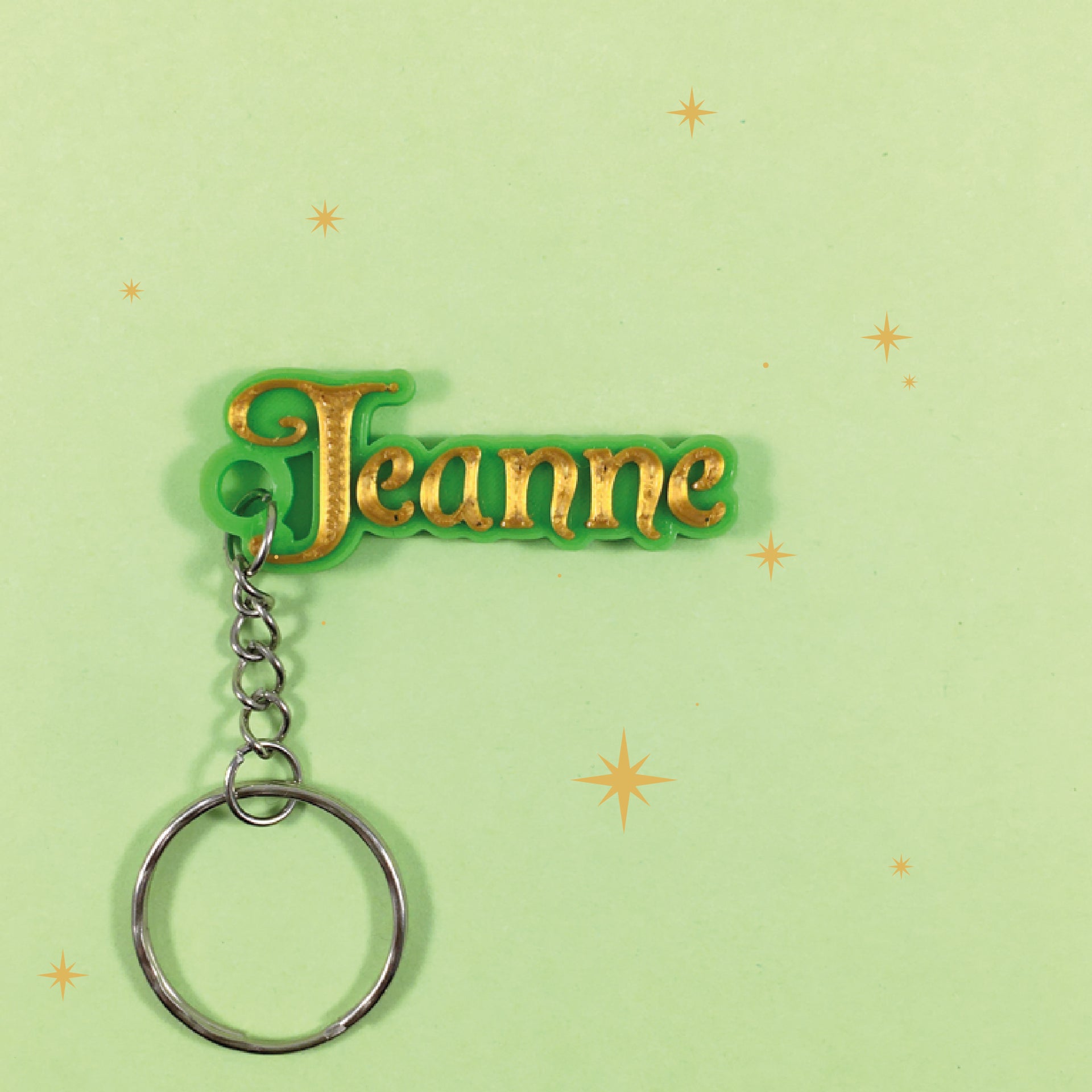 Porte-clés personnalisé style fée avec le prénom "Jeanne" en relief couleur or, monté sur un support vert. L'objet est posé sur un fond vert clair décoré de petites étoiles.