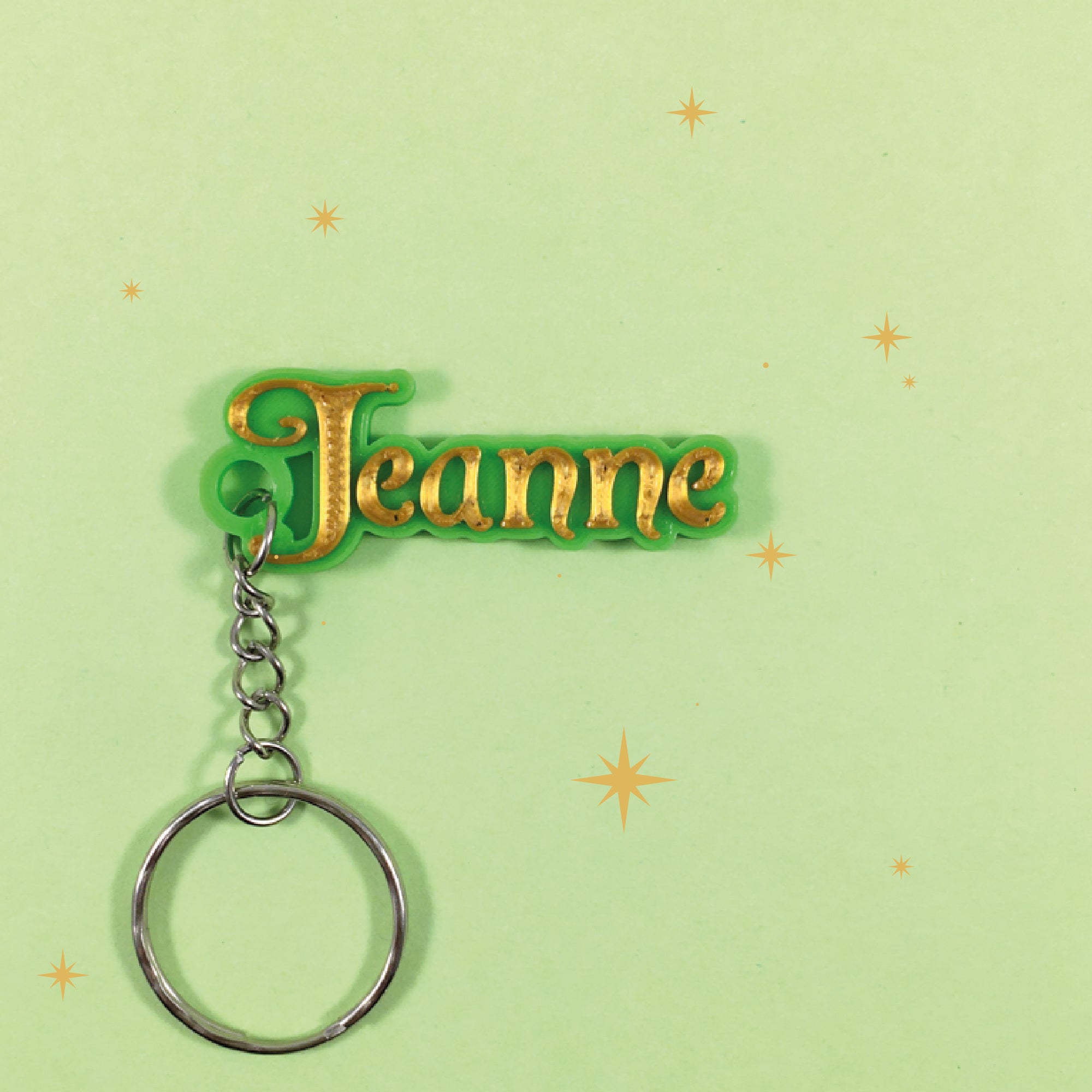 Porte-clés personnalisé style fée avec le prénom "Jeanne" en relief couleur or, monté sur un support vert. L'objet est posé sur un fond vert clair décoré de petites étoiles.