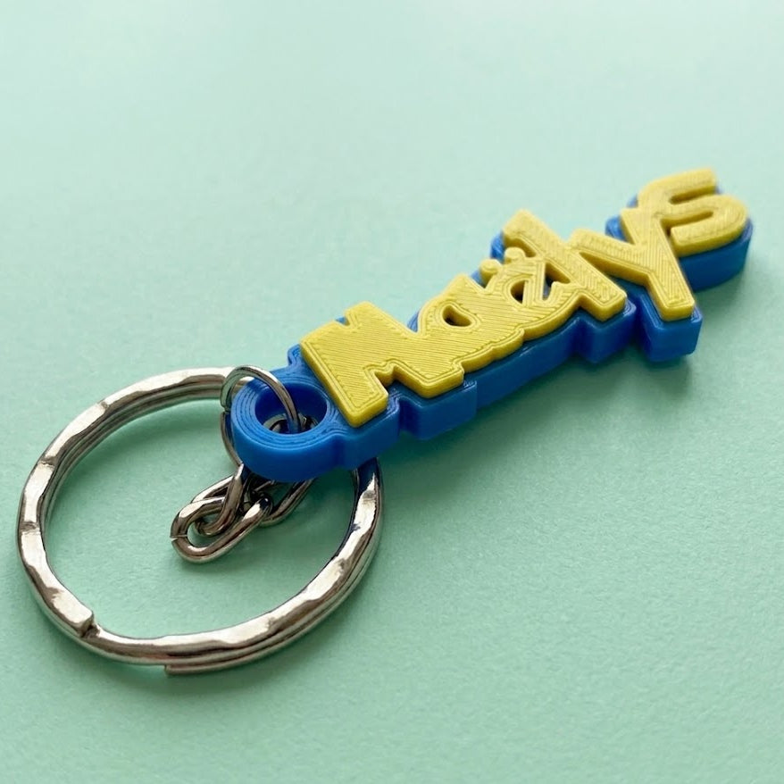 Porte-clés personnalisé imprimé en 3D au prénom Maëlys, avec texte en jaune vif sur une base bleu vif. L'attache est un anneau en métal argenté. L'objet repose sur un fond mat vert clair. Le design est d'inspiration "Dresseur d'Aventure Pokémon".