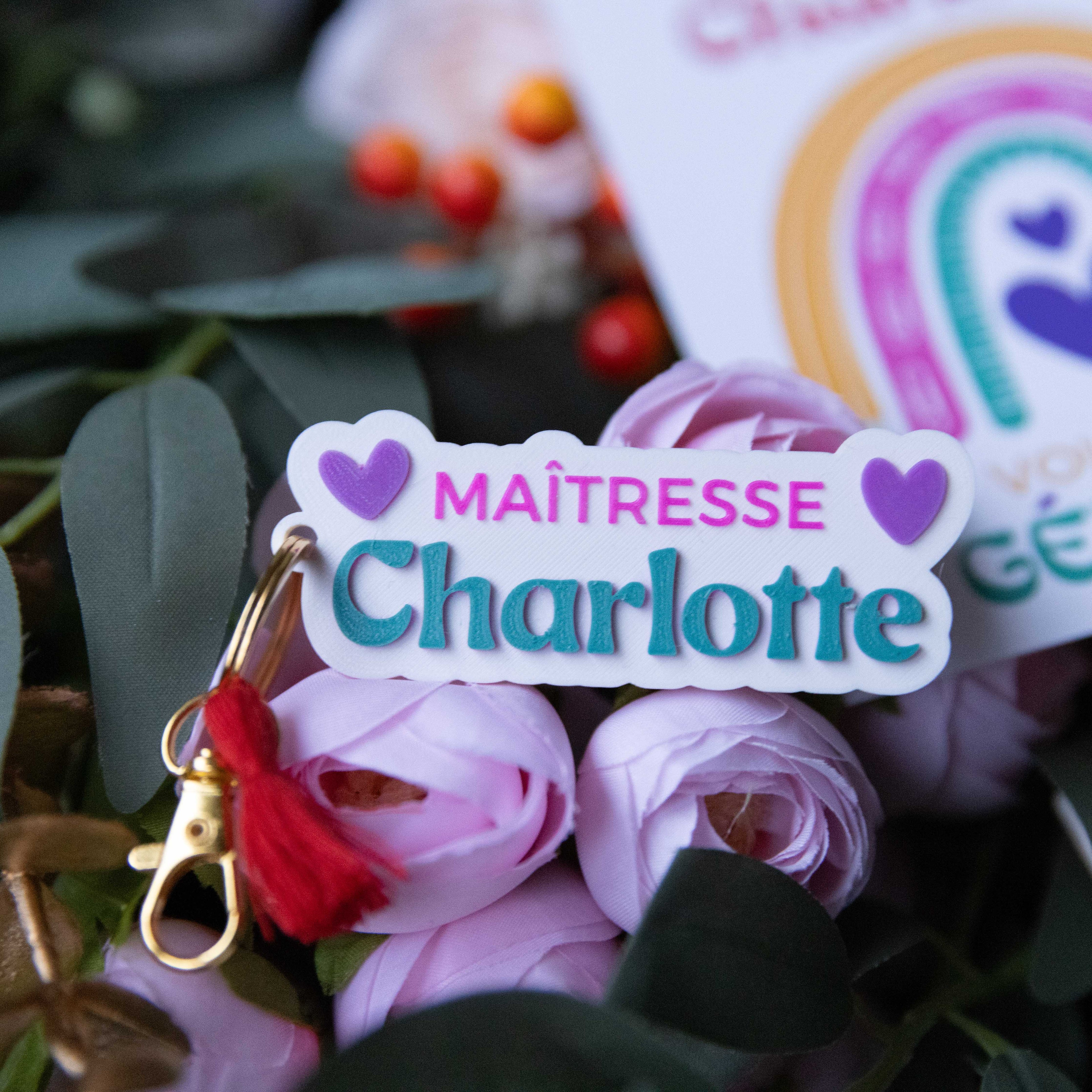 Gros plan d'un porte-clés personnalisé pour maîtresse au prénom 'Charlotte', avec texte rose et bleu et deux cœurs violets. L'attache dorée est décorée d'un petit pompon rouge, le tout posé sur des fleurs roses.
