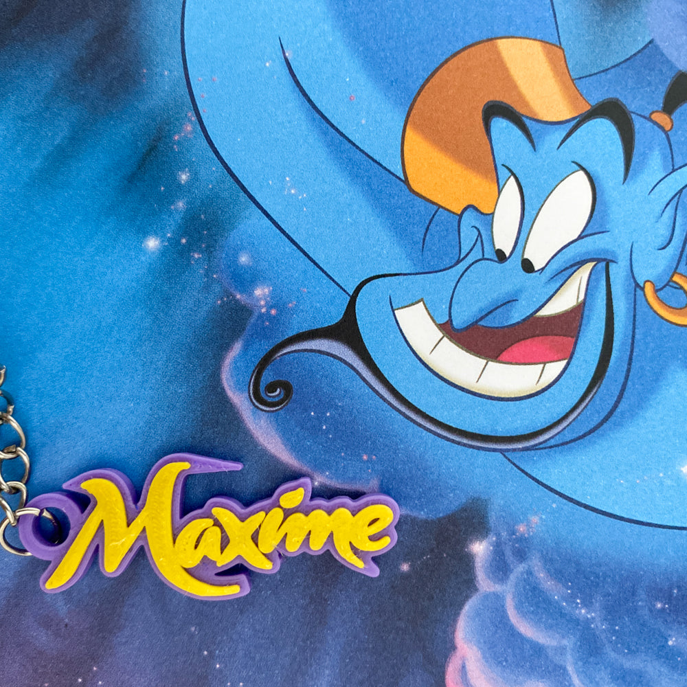Porte-clés personnalisé au prénom "Maxime" avec lettres or brillant sur support violet, posé sur une illustration du Génie d'Aladdin souriant de Disney sur fond de ciel étoilé bleu magique.