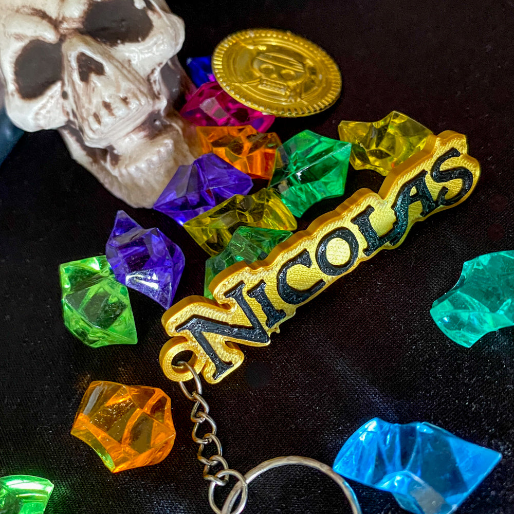 Porte-clés personnalisé 'NICOLAS' style pirate, avec le prénom en texte 3D noir en relief sur une base dorée. L'objet est posé sur un tissu noir au milieu d'un trésor composé de pierres précieuses colorées artificielles (vert, violet, orange), d'un crâne partiel et d'une pièce d'or de pirate.