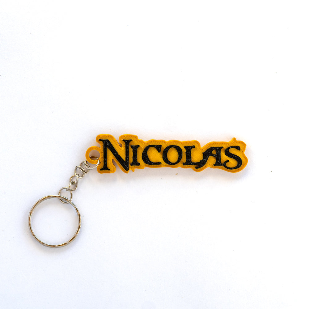 Porte-clés personnalisé avec le prénom NICOLAS, style pirate noir et or. Les lettres noires en relief sont sur une base or (jaune), avec une police stylisée pirate. Le porte-clés est attaché à une chaîne et un anneau brisé en métal argenté, sur fond blanc uni.