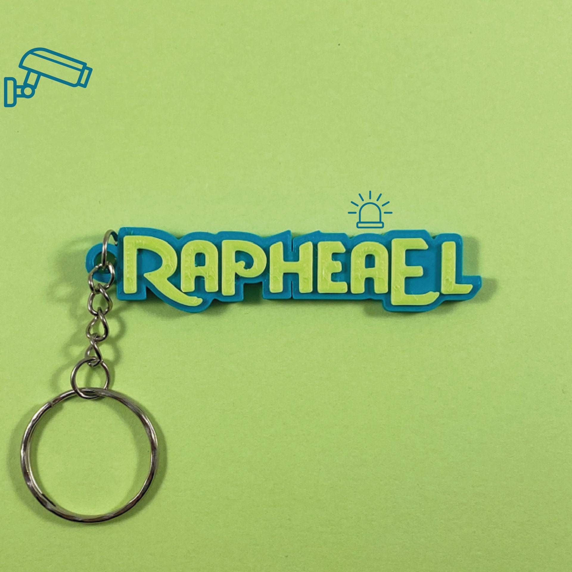 Porte-clés personnalisé au prénom Raphaël style Zootopie Métropole Animale, avec lettres vert pomme en relief sur support bleu. Le fond vert est décoré d'icônes de caméra de surveillance et d'une sirène de police.