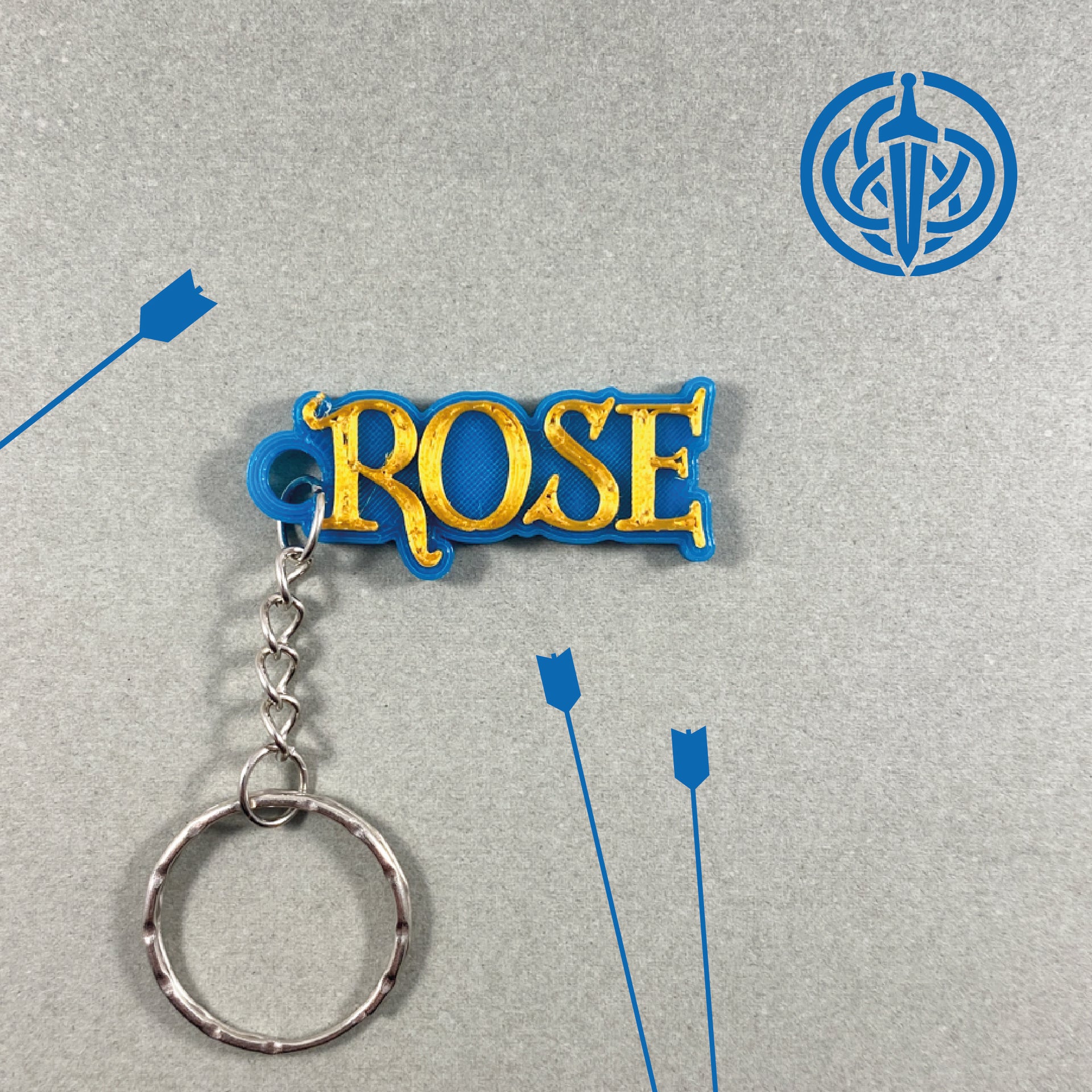Porte-clés personnalisé Rebelle au prénom ROSE, style médiéval fantastique avec lettres or en relief sur base bleue, sur fond gris avec illustrations de flèches et blason celtique avec épée bleus.