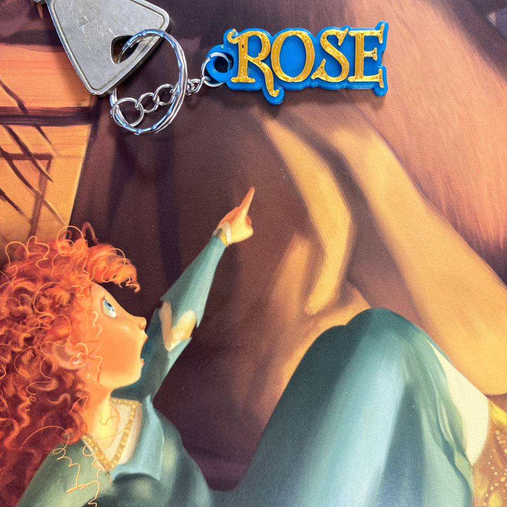 Porte-clés personnalisé au prénom ROSE, avec des lettres en relief couleur or sur une base bleue, posé sur une illustration thématique de Merida du film Disney Brave (Rebelle).