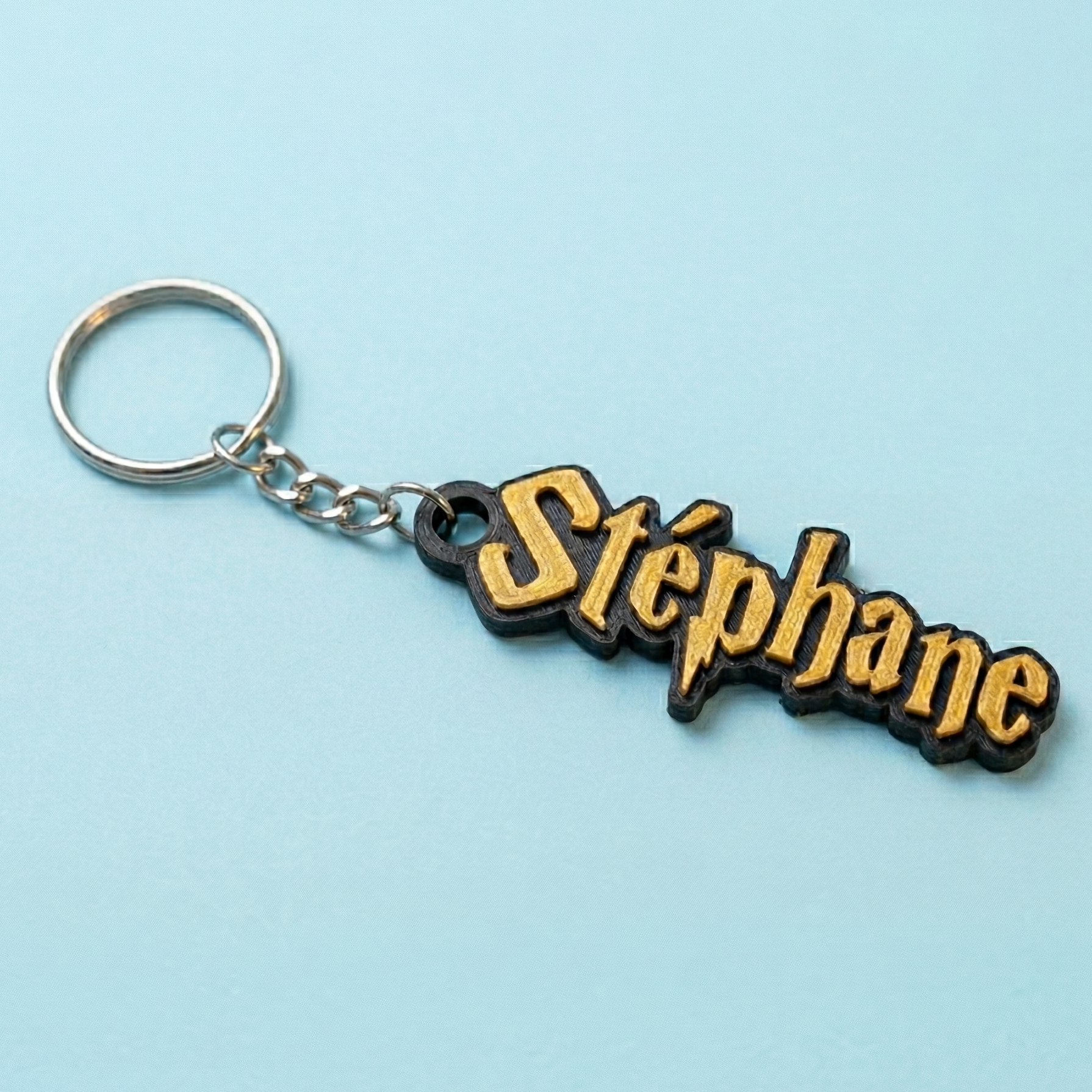 Porte-clés personnalisé au prénom Stéphane avec police d'écriture Harry Potter (éclair sur le P). Impression 3D avec lettres couleur or bronze en relief sur une base noire, présenté sur un fond bleu ciel.
