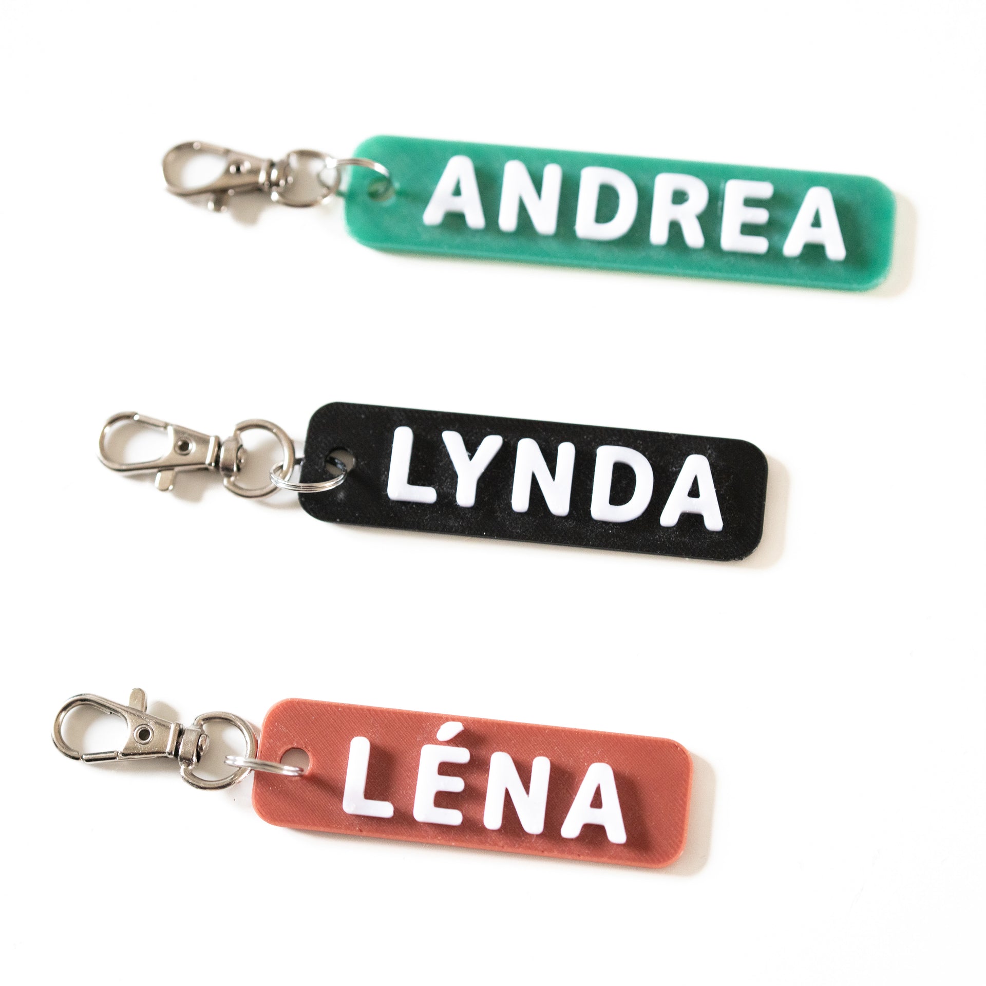 Vue de dessus de trois porte-clés personnalisés bicolores en relief 3D. Un porte-clé vert avec le prénom ANDREA, un noir avec LYNDA et un TOMETTE avec LÉNA. Lettres blanches en relief et fermoir mousqueton argenté sur fond blanc.