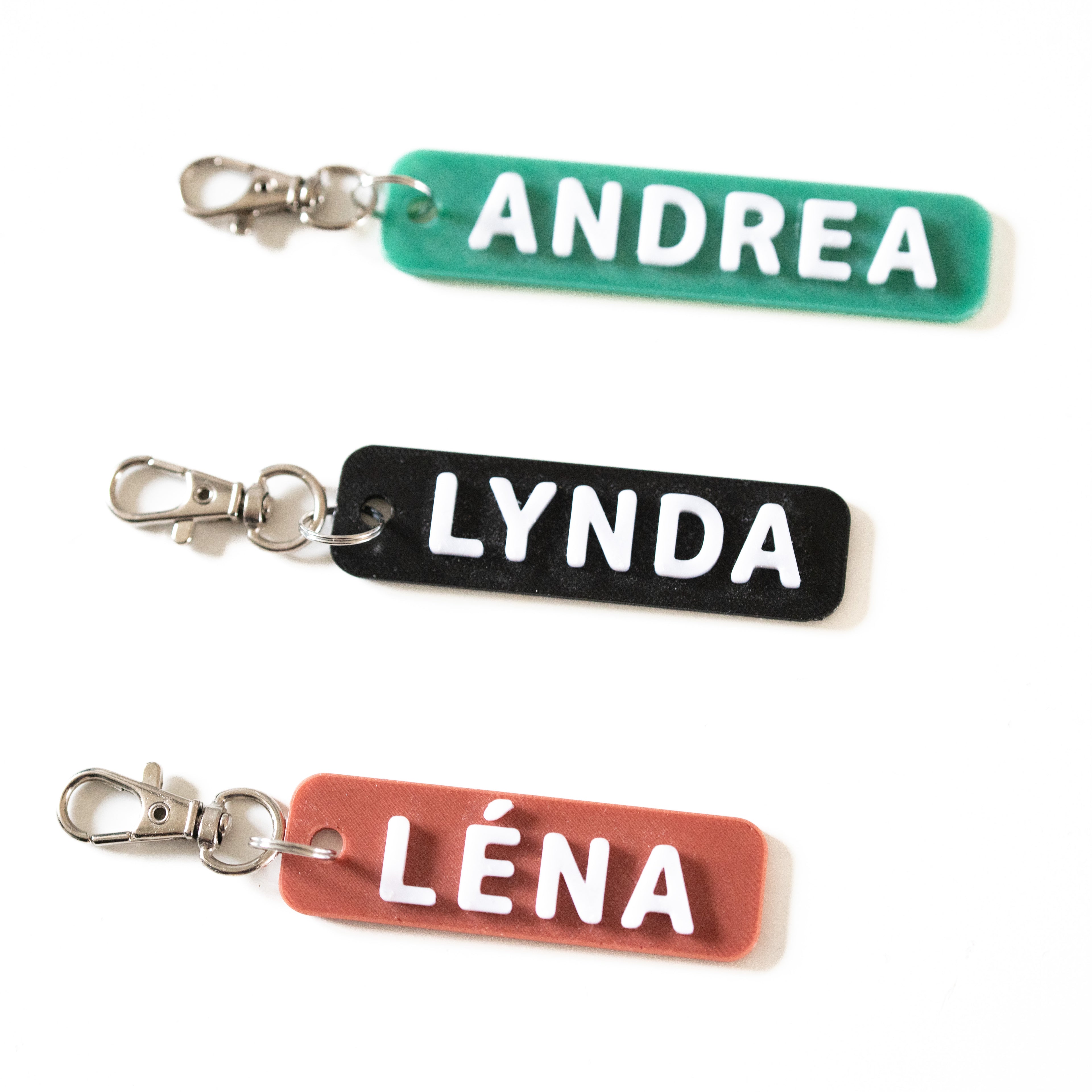 Vue de dessus de trois porte-clés personnalisés bicolores en relief 3D. Un porte-clé vert avec le prénom ANDREA, un noir avec LYNDA et un TOMETTE avec LÉNA. Lettres blanches en relief et fermoir mousqueton argenté sur fond blanc.