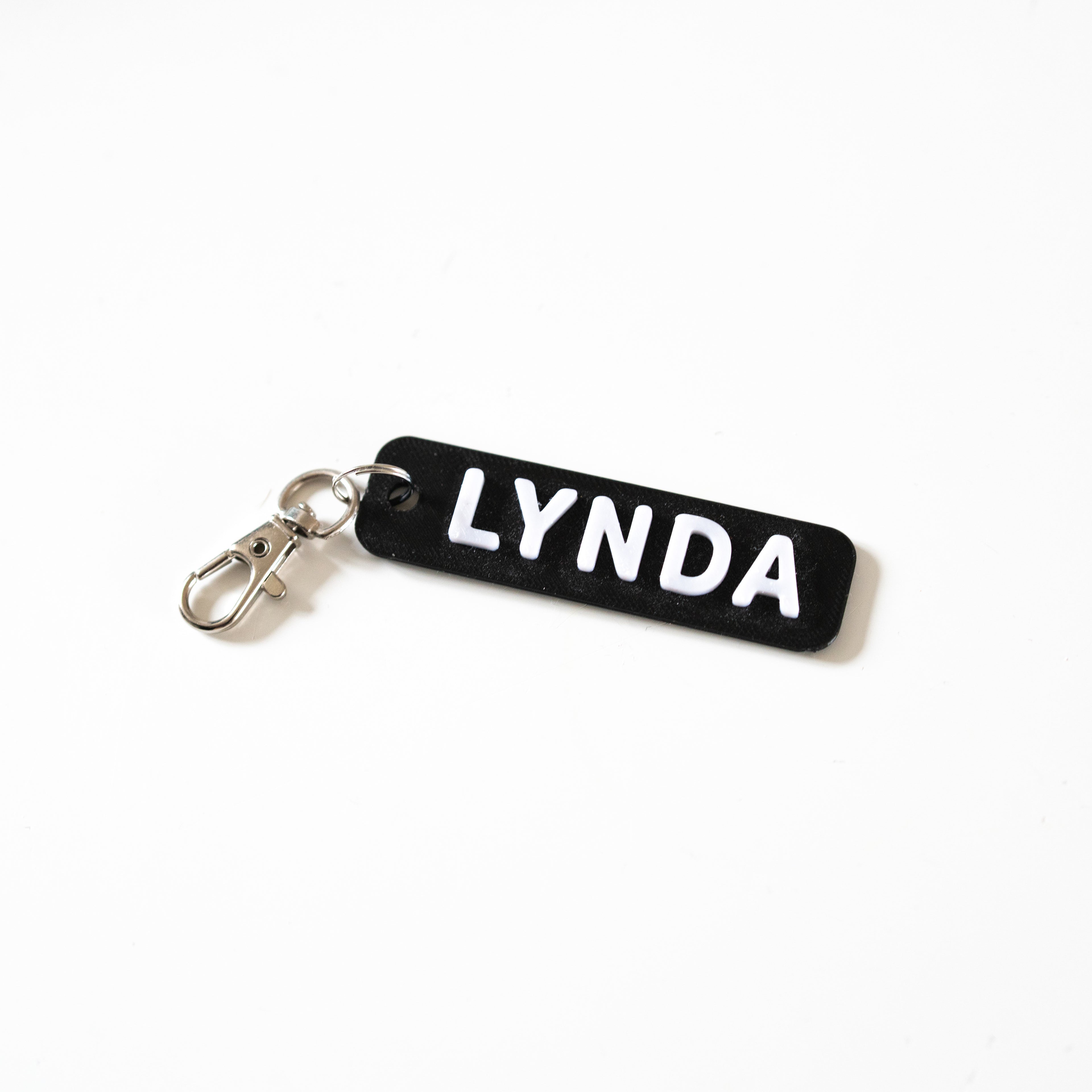 Porte-clé personnalisé en impression 3D avec le prénom LYNDA en relief blanc sur une base noire. Accessoire bicolore au design moderne avec attache mousqueton argentée sur fond blanc.