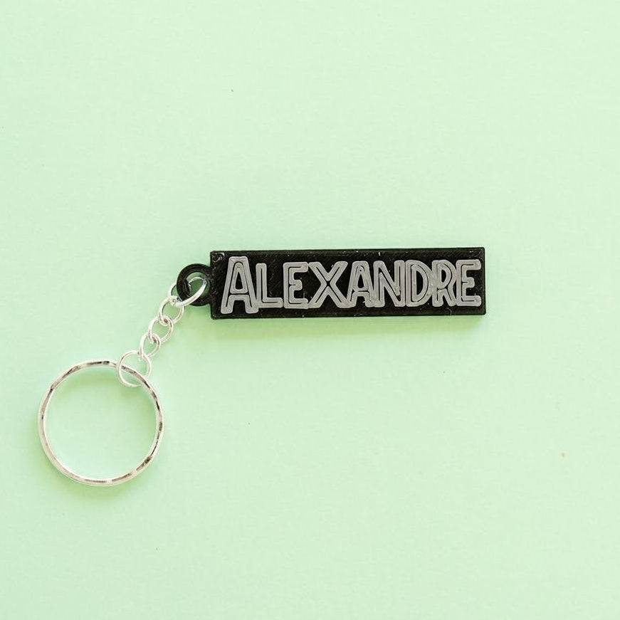 Porte-clés personnalisé noir et argent avec le nom ALEXANDRE en relief, dans une police de style Jurassic Park attaché à un anneau argenté sur un fond vert menthe uni.