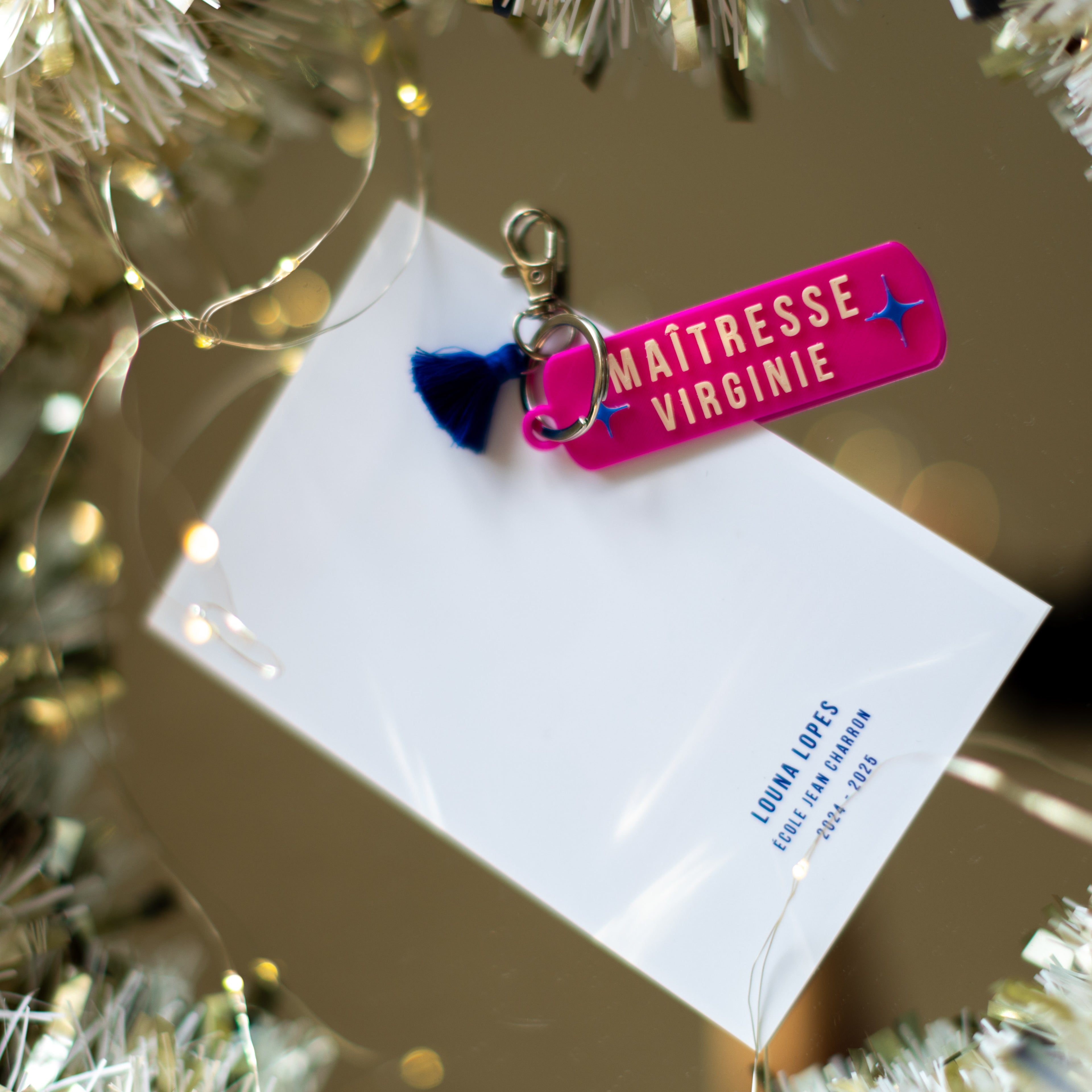 Kit cadeau pour maîtresse comprenant un porte-clés rose fuchsia et "MAÎTRESSE VIRGINIE" en blanc avec pompon bleu marine. Il est posé sur le verso de la carte personnalisée au nom de l'élève "LOUNA LOPES" de l'école Jean Charron pour l'année scolaire 2024-2025. Décoration festive avec guirlande argentée et lumières de Noël