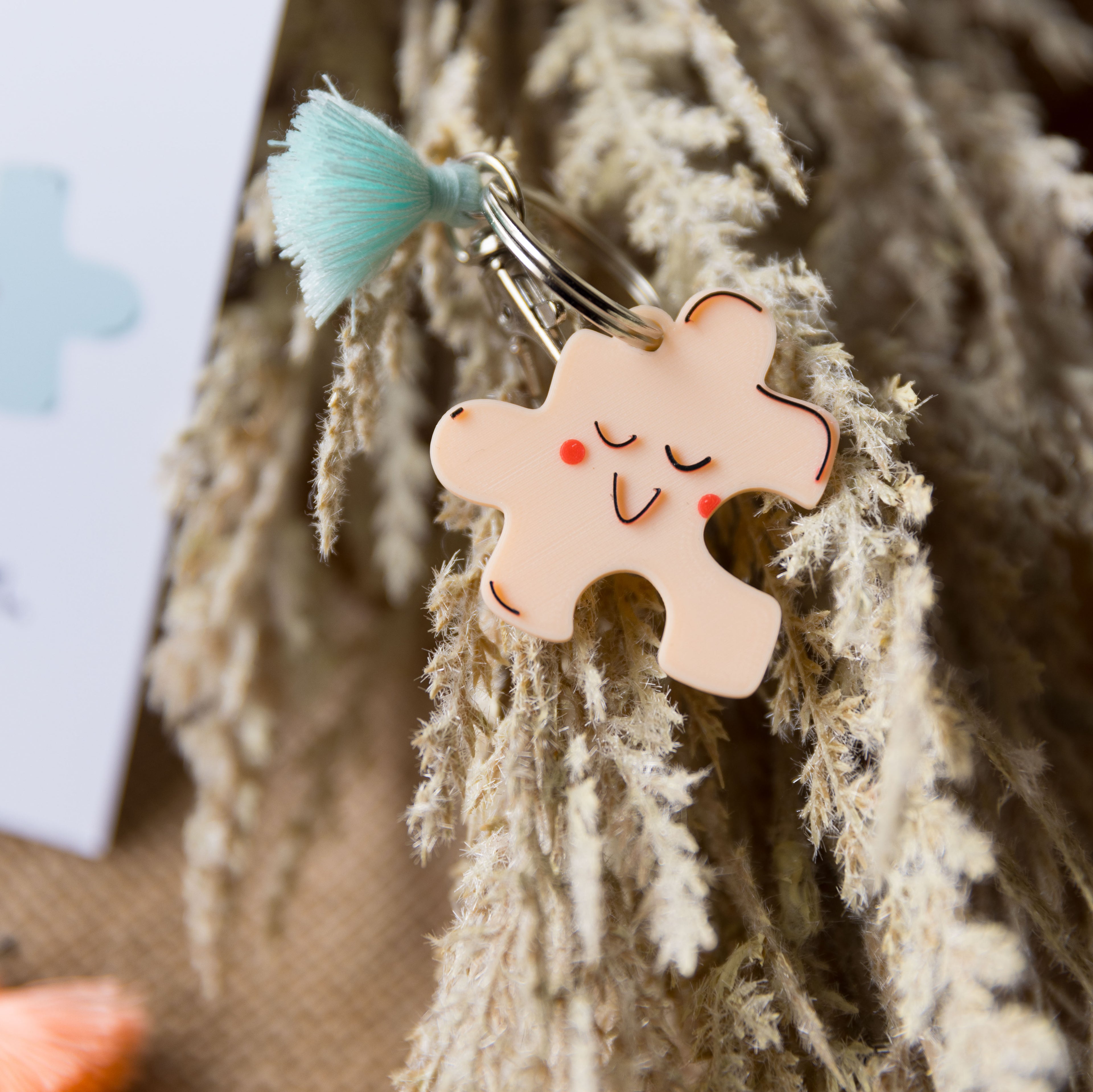 Gros plan d'un porte-clés puzzle réalisé en impression 3D PLA de couleur pêche, laissant apparaître une texture finement striée. Le visage "sleepy" est imprimé avec des détails rouges. Il est accompagné d'un pompon vert menthe, le tout posé sur un décor naturel d'herbes séchées.