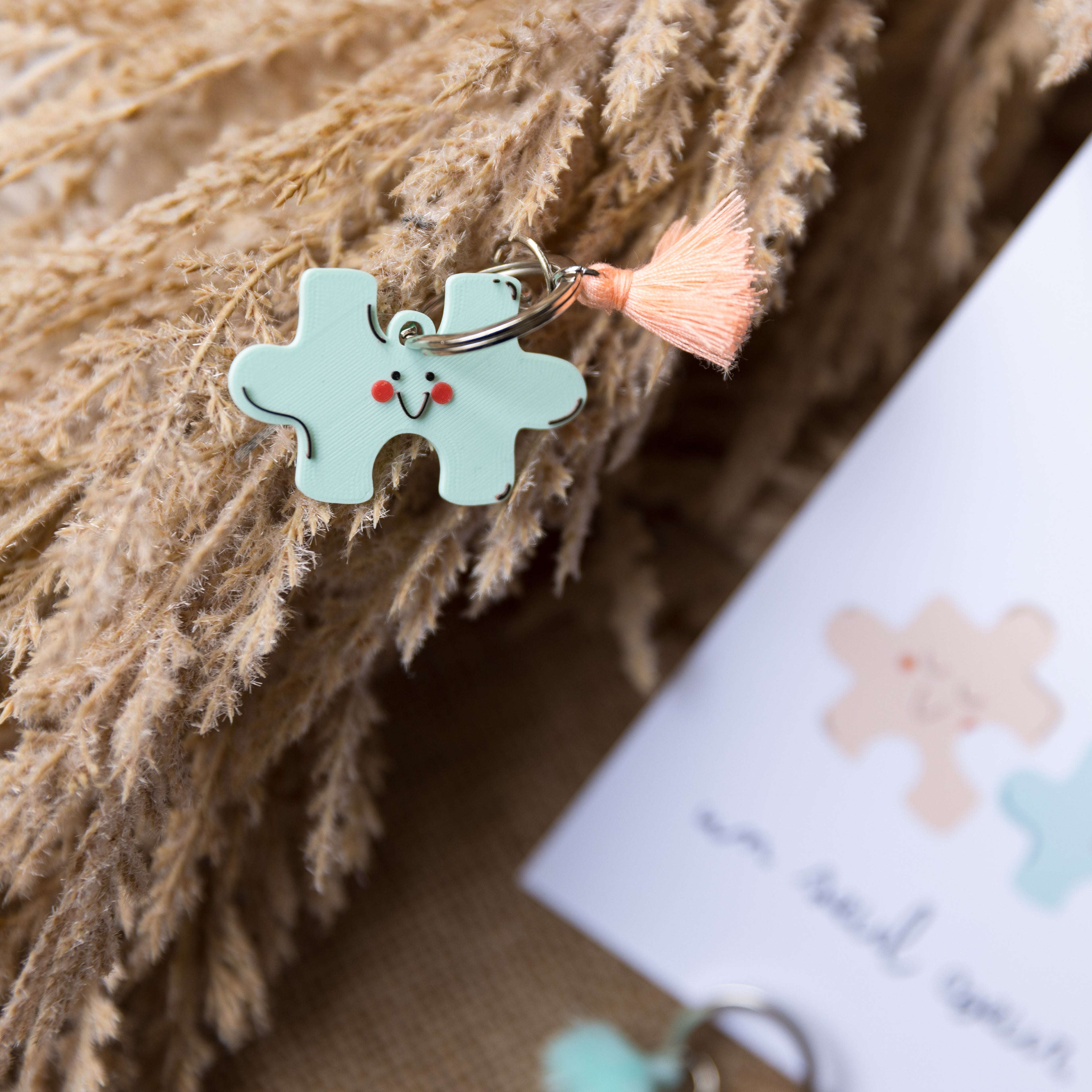 Gros plan d'un porte-clés en forme de pièce de puzzle vert d'eau clair avec un visage souriant noir et des petites joues rouges, un pompon corail pêche et un anneau argenté, posé sur des herbes séchées avec une carte floue.