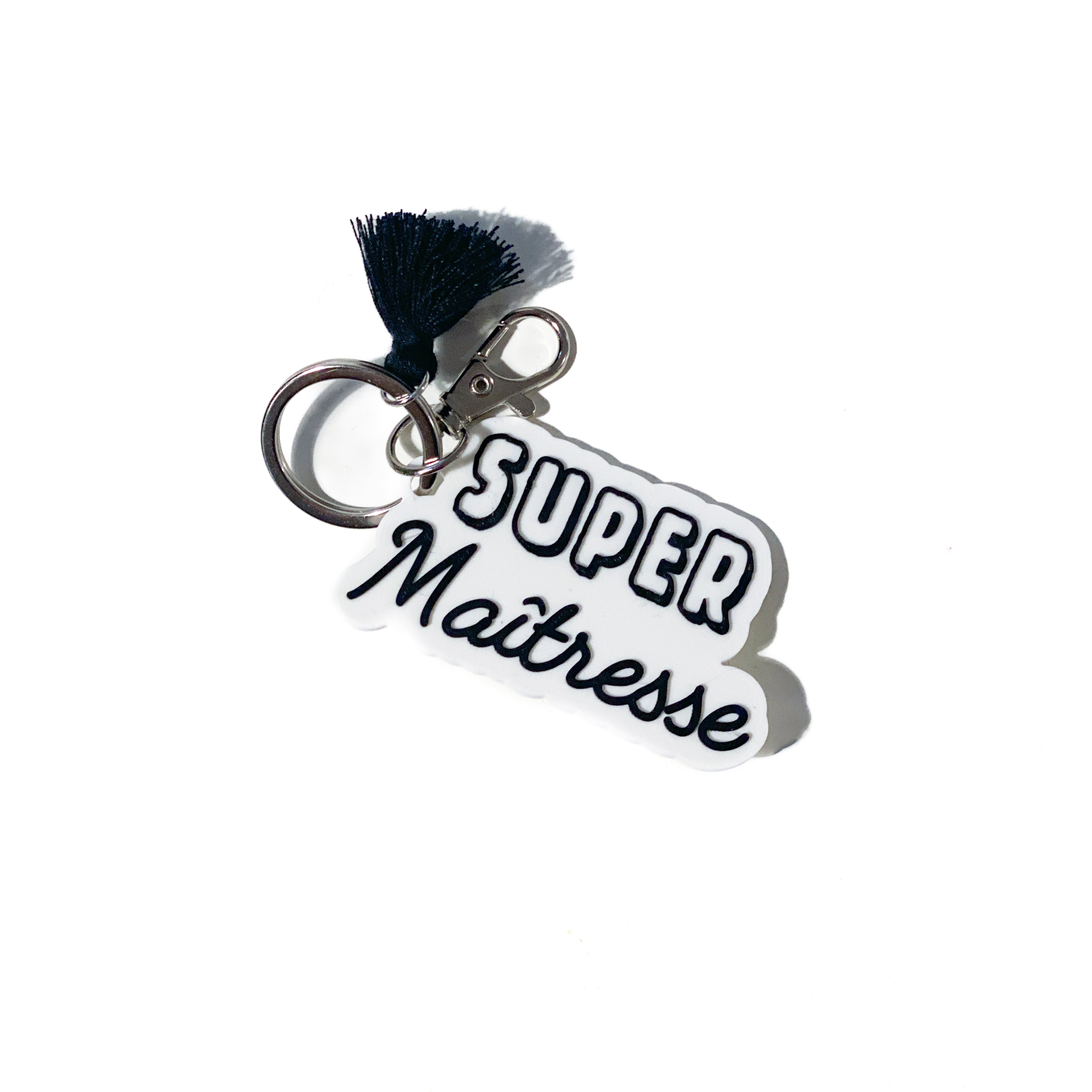Porte-clés blanc réalisé en impression 3D avec l'inscription en relief noir "SUPER Maîtresse". Le mot "SUPER" est en lettres capitales arrondies et "Maîtresse" en écriture manuscrite élégante. L'accessoire inclut un anneau argenté, un fermoir mousqueton et un pompon décoratif en fil noir, présenté sur un fond blanc uni.