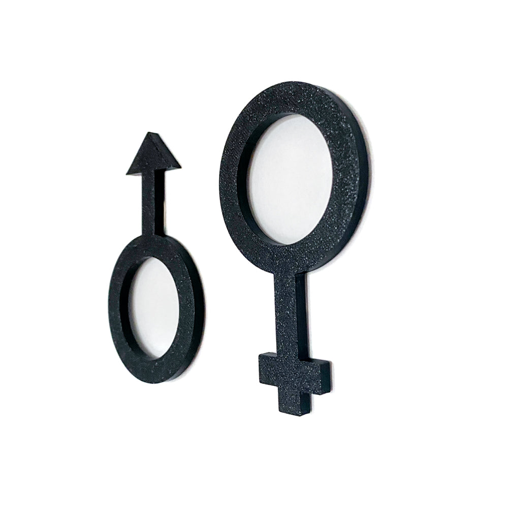Gros plan en perspective de deux pictogrammes noirs de toilettes, symbole masculin et symbole féminin en relief, imprimés en 3D pour signalétique de porte.