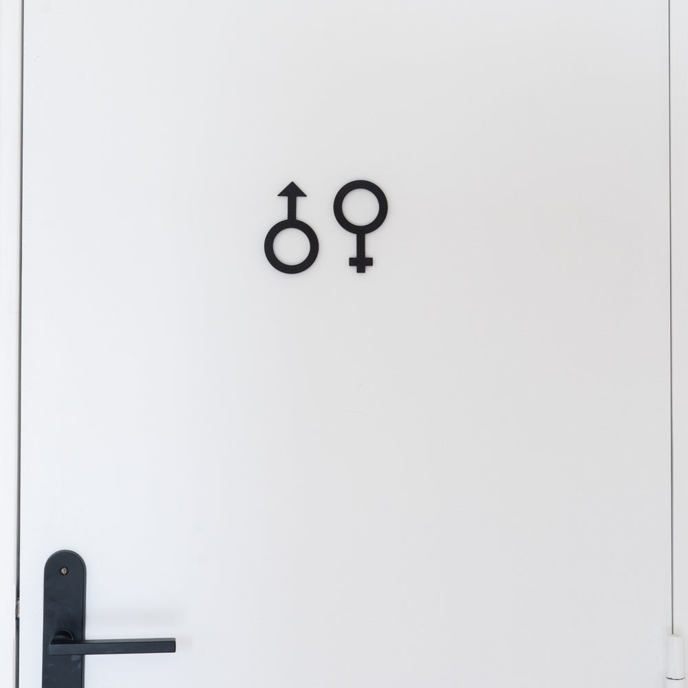 Porte blanche de toilettes avec deux pictogrammes noirs homme et femme en relief au centre et poignée noire en bas à gauche, signalétique WC mixte au design minimaliste.