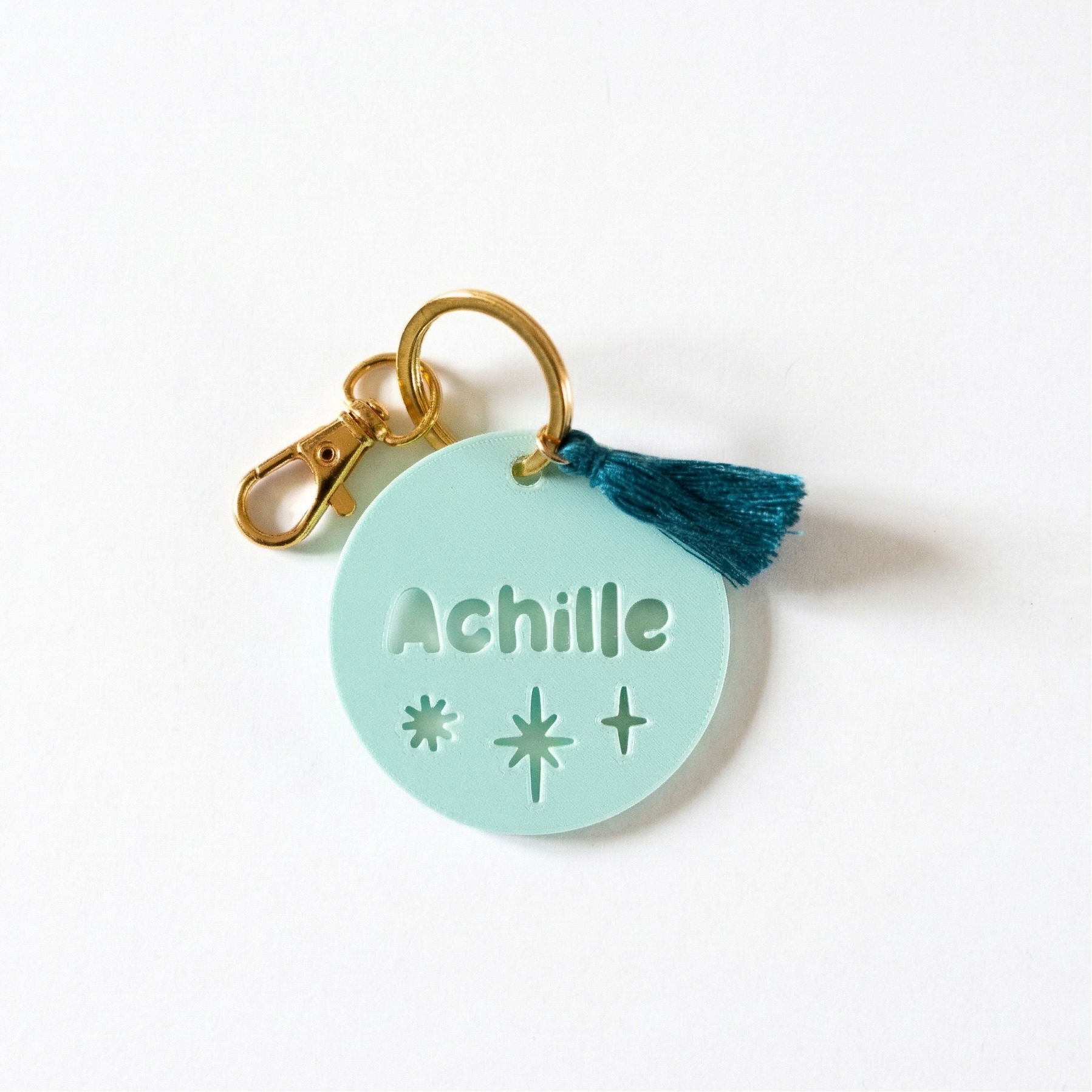Étiquette de sac personnalisée Achille en impression 3D vert d’eau avec motifs étoiles, mousqueton doré brillant et pompon soyeux vert canard sur fond bleu.