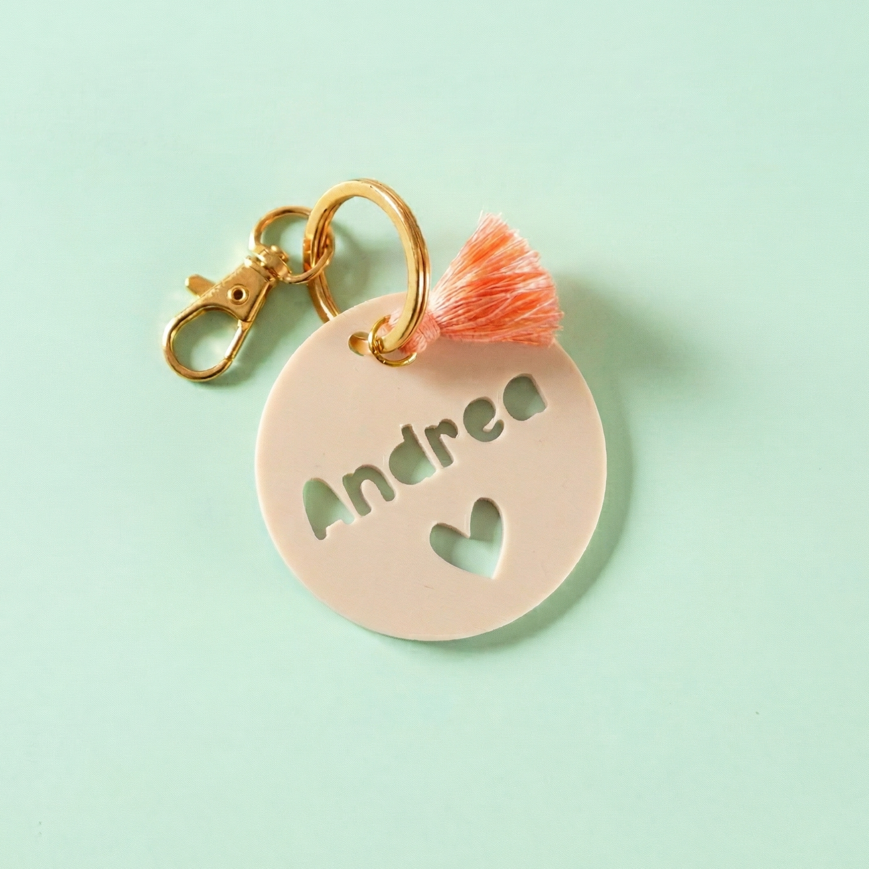 Étiquette de sac personnalisée Andrea en impression 3D beige avec motif cœur découpé, mousqueton doré brillant et pompon corail orangé sur fond vert pâle.