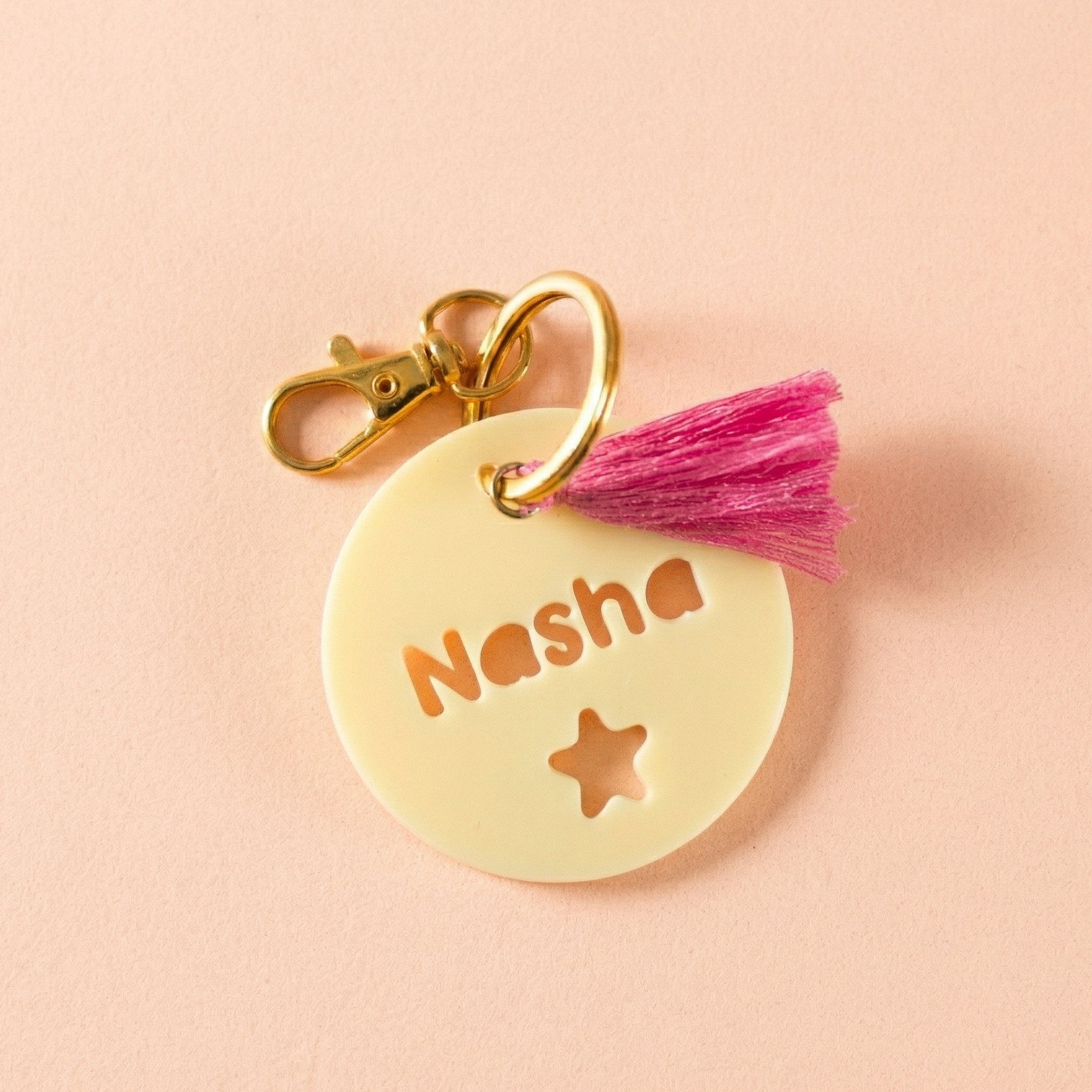 Vue en gros plan d'un médaillon de sac personnalisé en impression 3D jaune pastel. Le nom 'Nasha' et une étoile sont découpés avec précision. Le médaillon est attaché à une attache dorée comprenant un anneau et un mousqueton, et orné d'un pompon rose vif. Fond pêche.