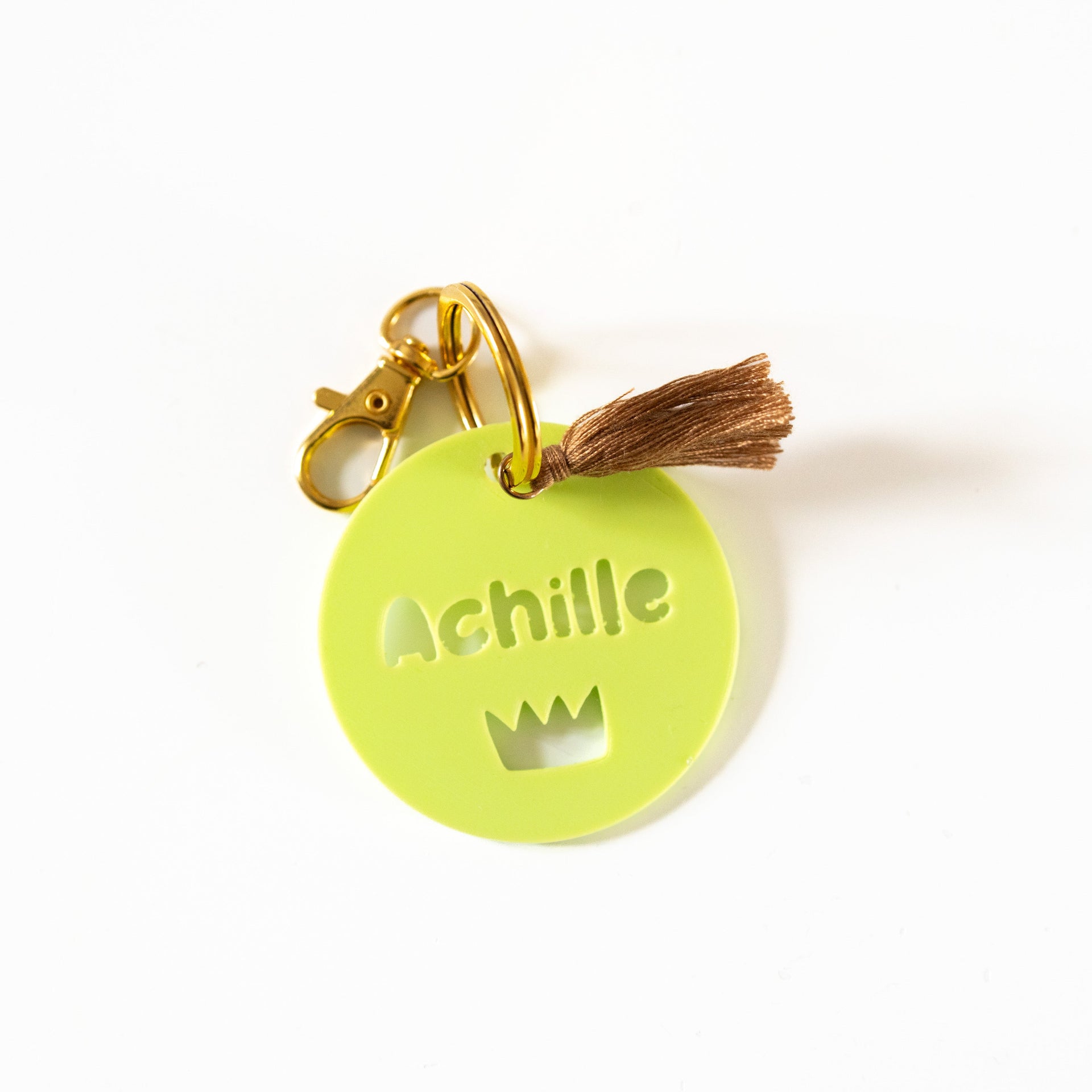 Étiquette de sac personnalisée Achille en impression 3D vert pistache lumineux avec motif couronne découpé, mousqueton doré et pompon brun soyeux sur fond blanc.