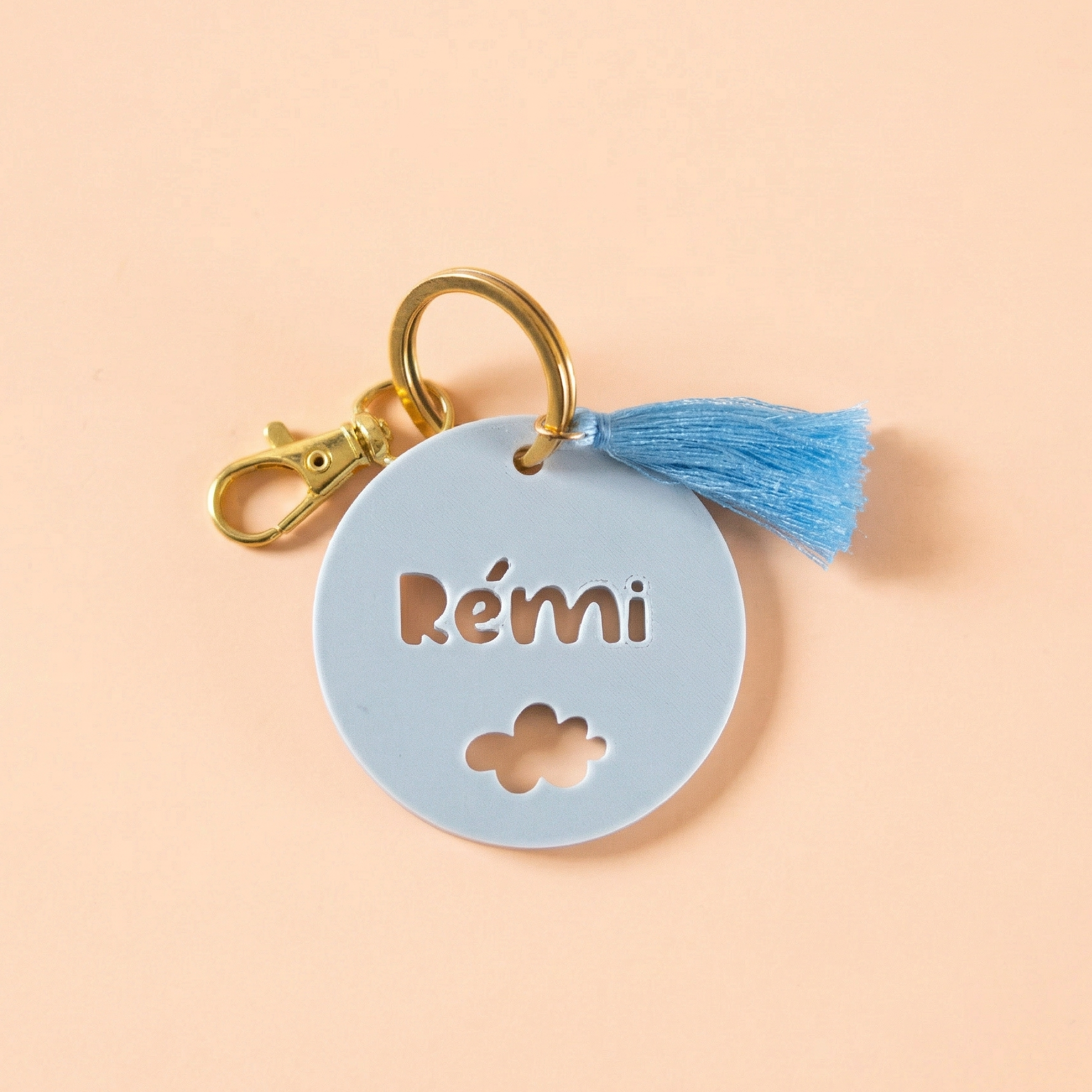 Étiquette de sac personnalisée Rémi en impression 3D gris bleuté clair avec motif nuage découpé, mousqueton doré brillant et pompon bleu soyeux sur fond rose pêche.