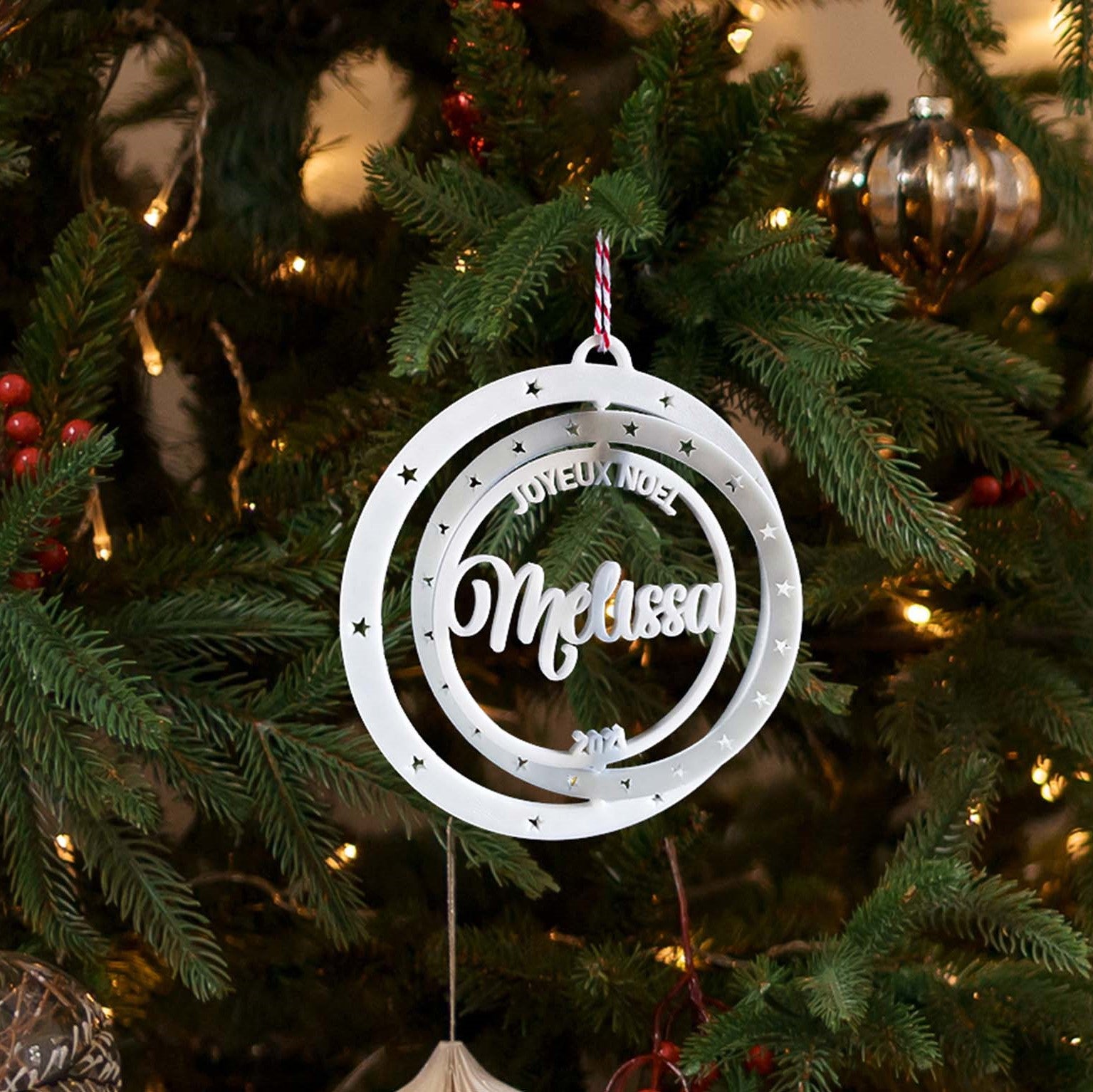 Boule de Noël personnalisable suspendue sur un sapin : double cercle ajouré avec petites étoiles, texte “Joyeux Noël”, prénom au centre (ex. “Melissa”) et année en bas, sur fond de branches de sapin illuminées.