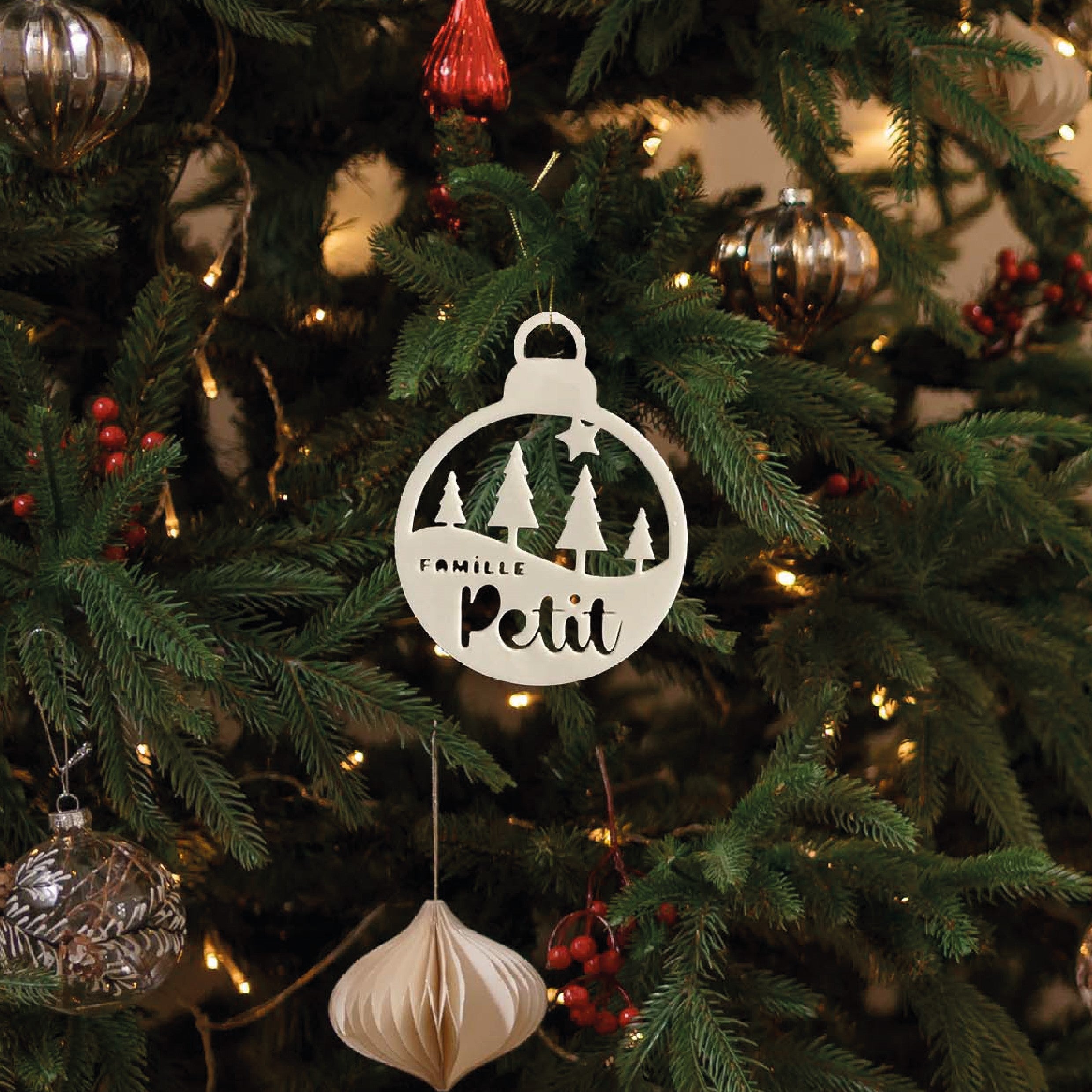 Boule de Noël personnalisable “La forêt familiale” en décoration de sapin : ornements découpés avec silhouettes de sapins et texte “Famille” + prénom (ex. “Petit”), suspendu sur un sapin illuminé.