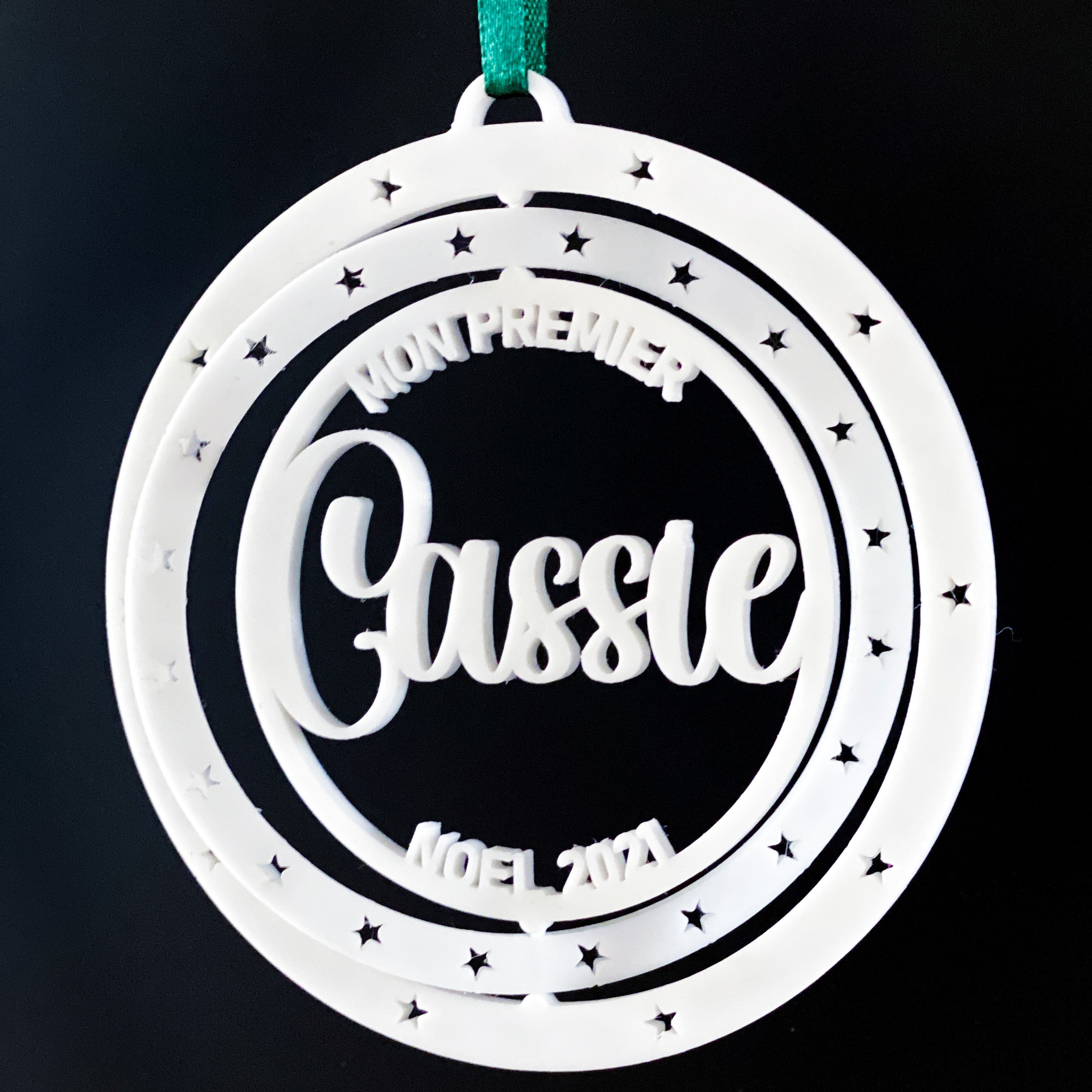 Boule de Noël personnalisable “Mon premier Noël” : décoration blanche en double cercle ajouré avec étoiles, prénom en relief au centre (ex. “Cassie”) et mention “Noël 2024” en bas, suspendue par un ruban vert sur fond noir.
