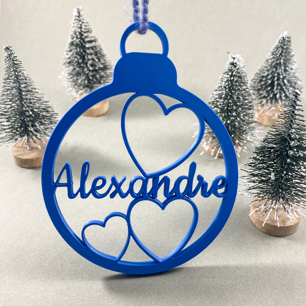 Boule de Noël personnalisable bleue avec prénom “Alexandre” et trois cœurs ajourés, suspendue par une ficelle, mise en scène devant de petits sapins décoratifs enneigés sur fond clair.