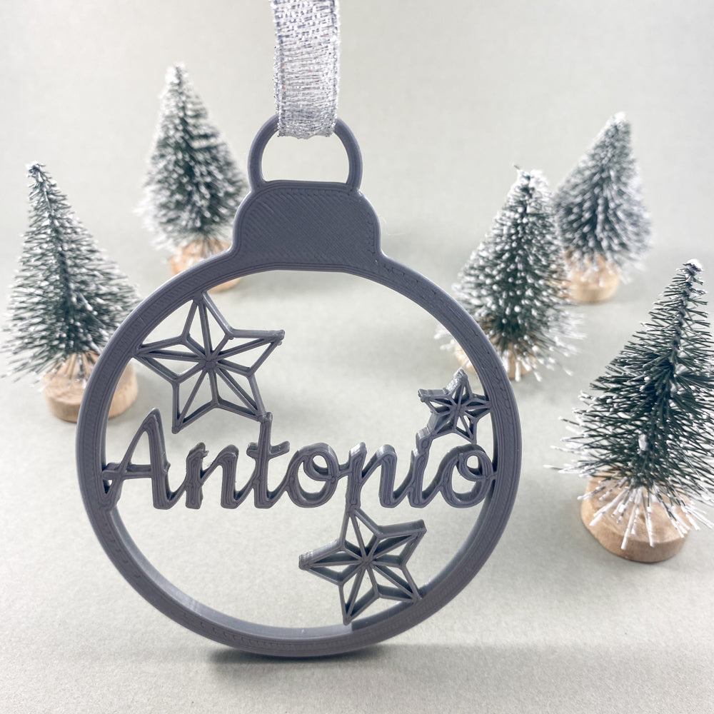 Boule de Noël personnalisable argent avec prénom “Antonio” et trois étoiles ajourées, suspendue par un ruban argenté, mise en scène devant de petits sapins décoratifs enneigés sur fond clair.