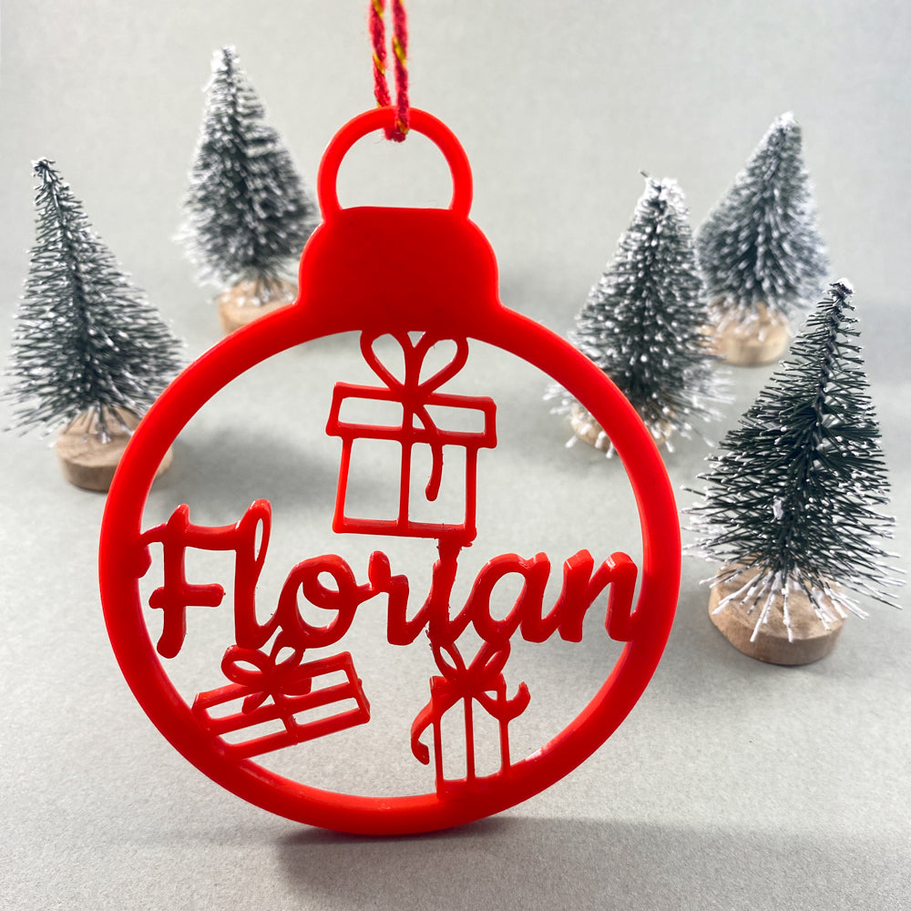 Boule de Noël personnalisable rouge avec prénom “Florian”  et motifs de cadeaux, suspendue par une ficelle rouge, mise en scène devant de petits sapins décoratifs enneigés sur fond clair.