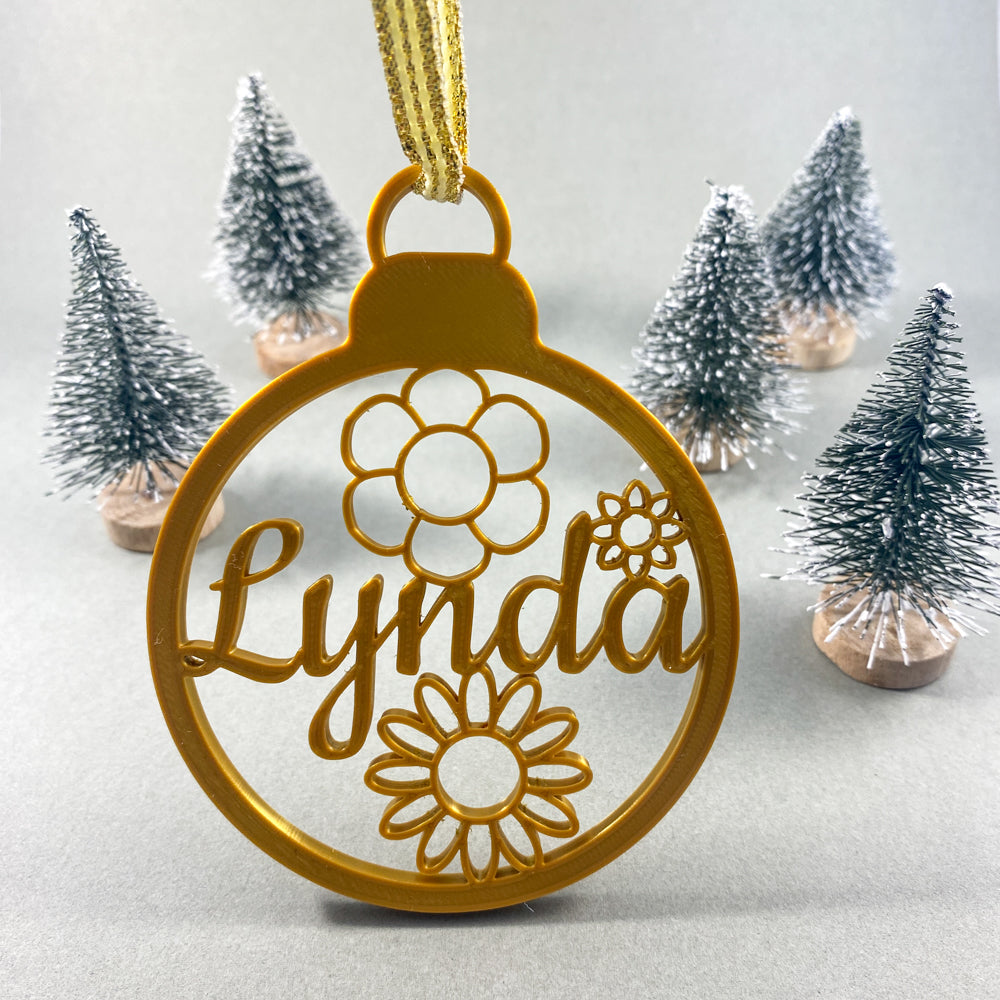 Boule de Noël personnalisable couleur or avec prénom “Lynda” découpé et motifs de fleurs à l’intérieur, suspendue par un ruban doré, mise en scène devant de petits sapins décoratifs enneigés sur fond clair.
