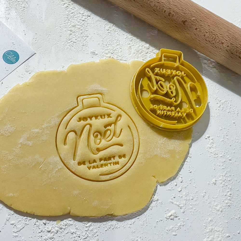 Emporte-pièce en forme de boule de Noël personnalisé insérant le prénom « Valentin », posé sur une pâte à biscuits étalée, avec l’empreinte du sablé bien visible et un rouleau à pâtisserie en arrière-plan.