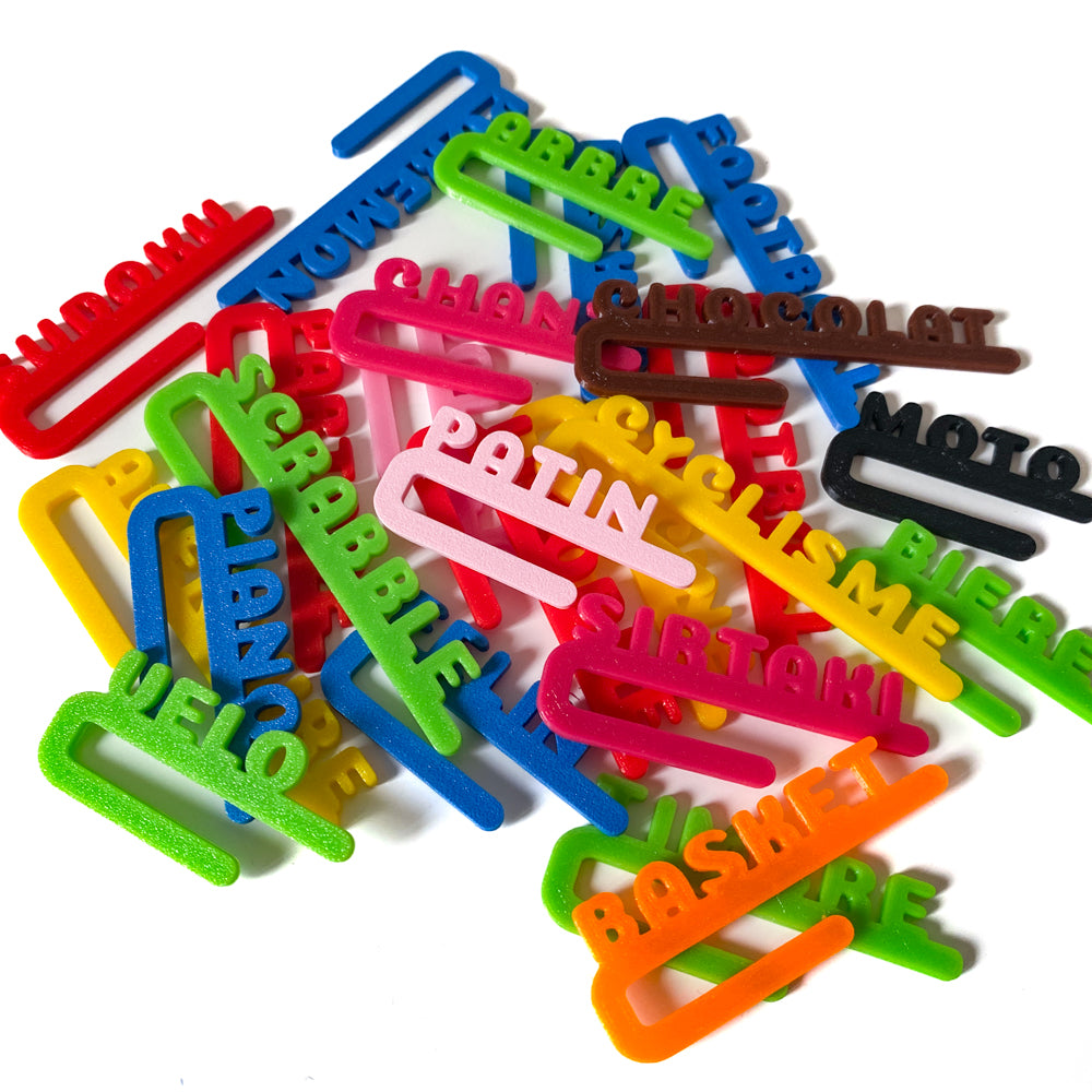 Grande variété de clips marque-verres Linéal en PLA, dispersés en vrac : chaque pièce est une barre fine avec des lettres droites formant des prénoms ou mots comme “Scrabble”, “Basket”, “Patin”, “Piano”. Les couleurs sont très vives (vert, bleu, rose, jaune, orange, rouge, noir). Les pièces se chevauchent sur un fond blanc.