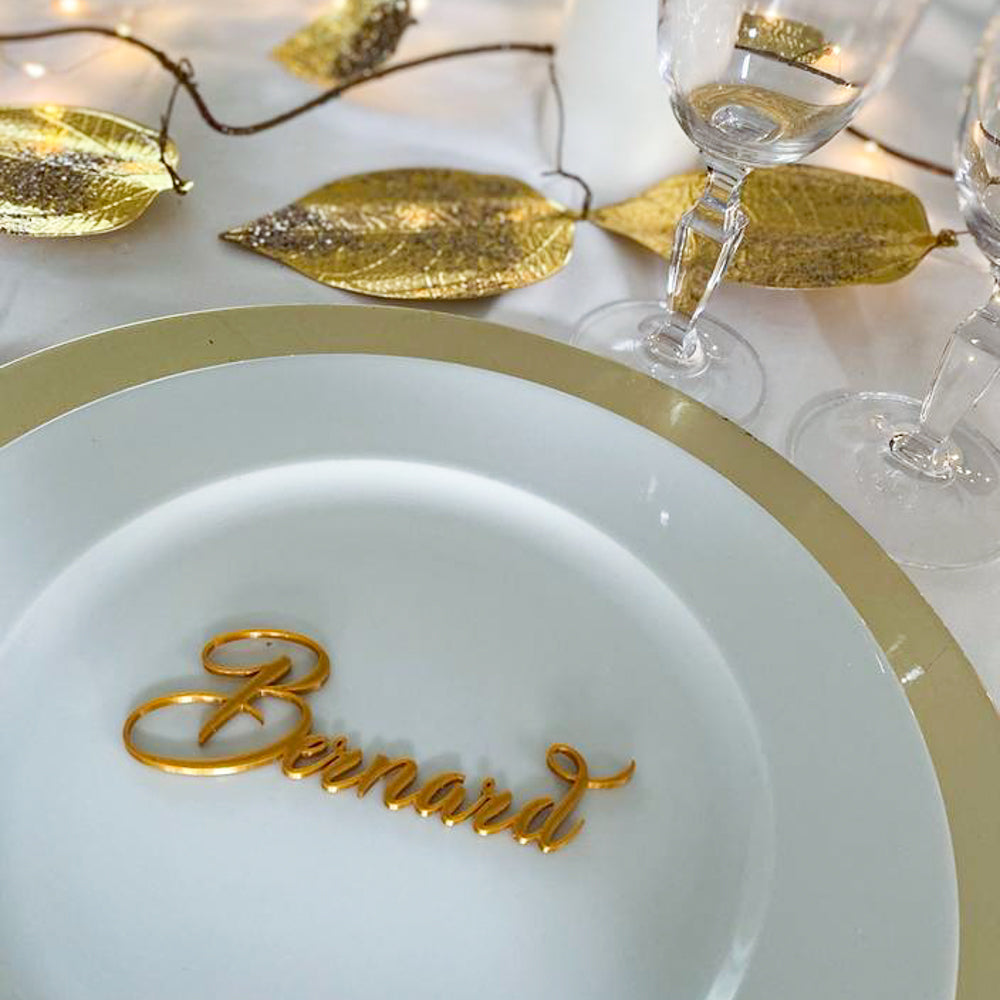 Marque-place personnalisé en écriture calligraphique dorée posé sur une assiette blanche et or, idéal pour une table élégante de fête ou de réception.