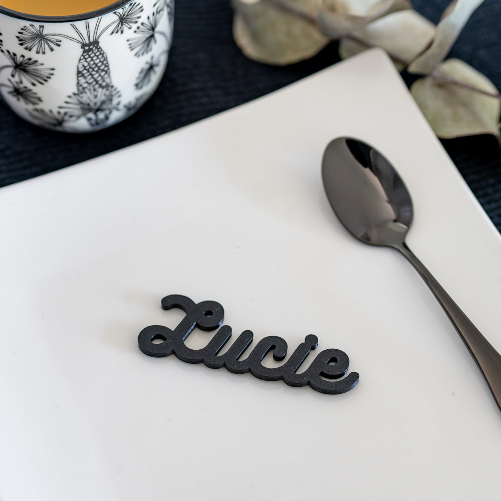 Marque-place cursive personnalise avec le prenom “Lucie” pose sur une assiette blanche, style moderne et lisible.
