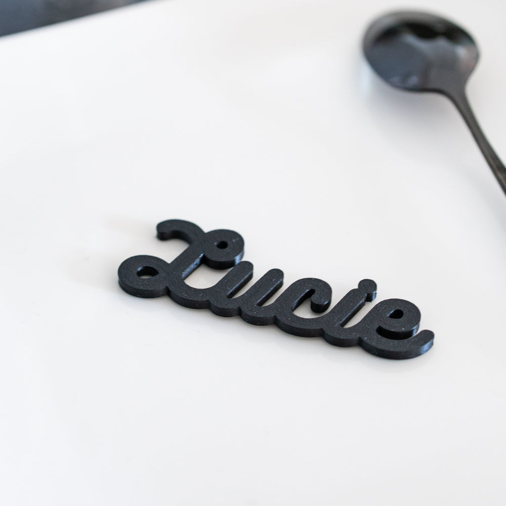 Zoom sur le marque-place cursive noir avec prénom « Lucie », imprime en PLA, présenté sur une assiette blanche.