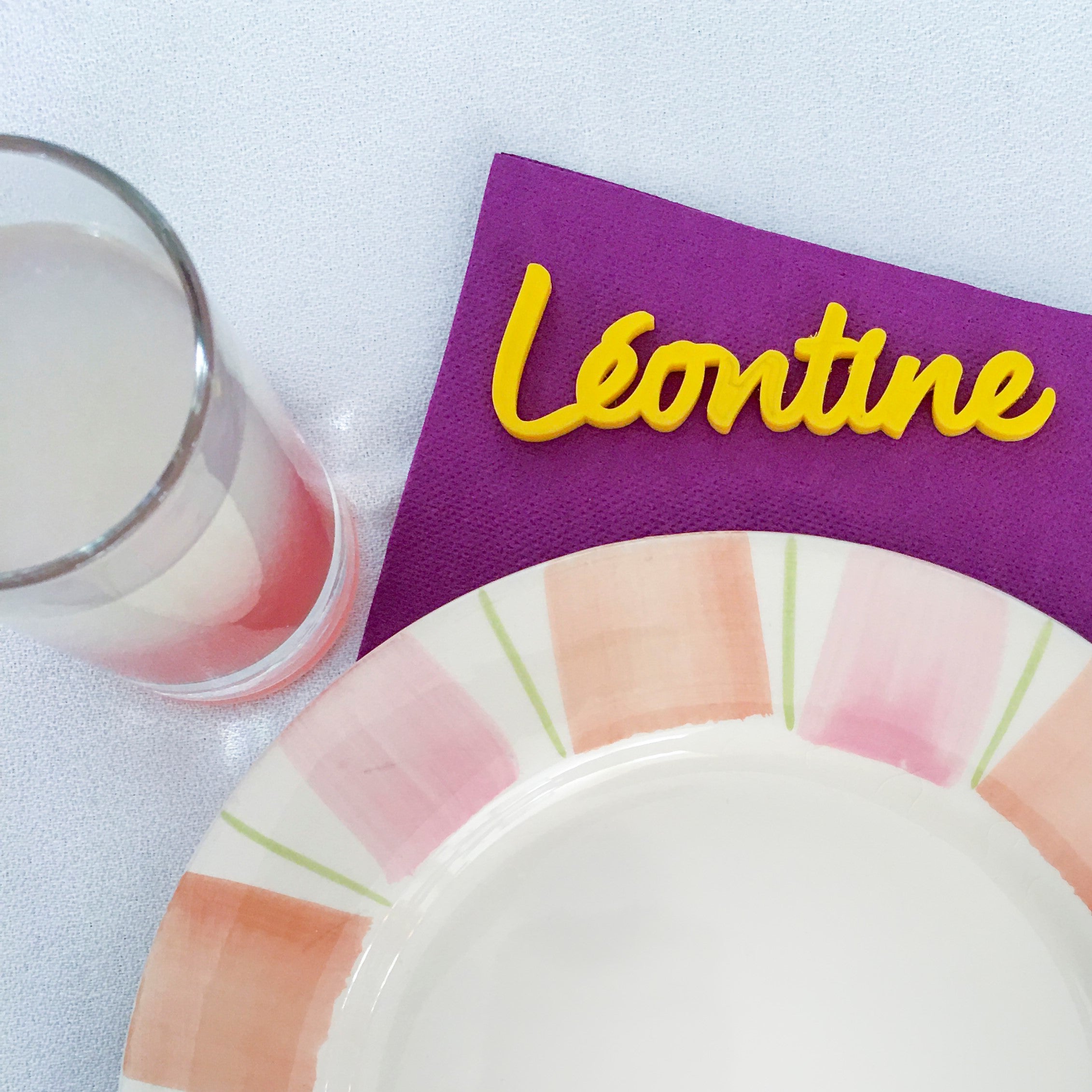 Marque-place jaune en impression 3D avec le prénom Léontine, posé sur une serviette violette aux côtés d’une assiette pastel pour une table joyeuse et raffinée.