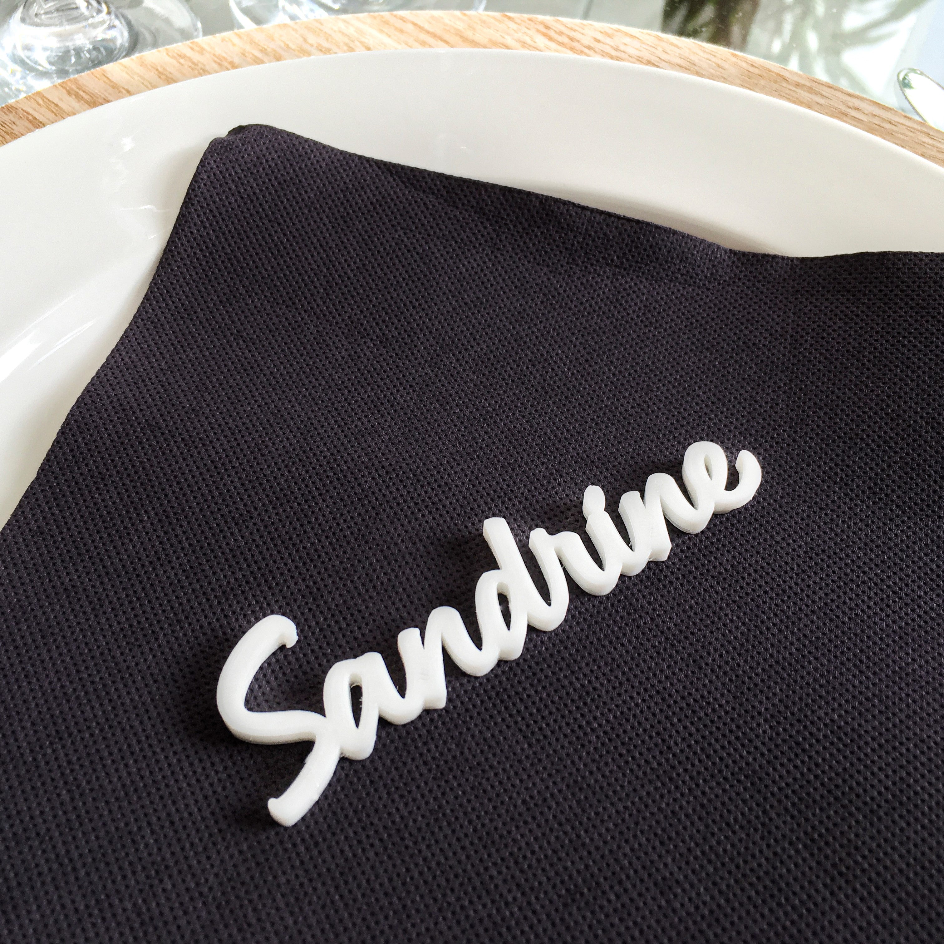 Marque-place blanc en impression 3D avec le prénom Sandrine, posé sur une serviette noire pour une décoration de table élégante et raffinée.