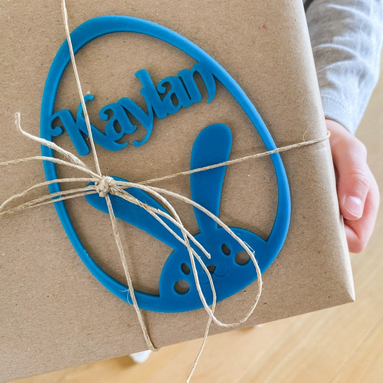 Marque-place de Pâques en forme d’œuf avec lapin et prénom « Kaylan » intégré, utilisé comme étiquette décorative sur un paquet cadeau kraft.