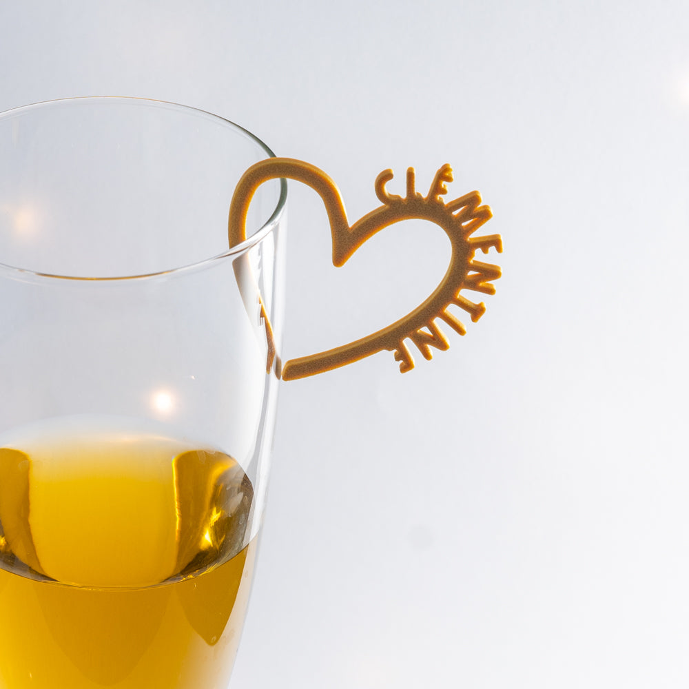 Photo d’un verre à pied rempli d’une boisson dorée, décoré d’un marque-verre en forme de cœur couleur doré, personnalisé avec le prénom « Clémentine ». Le cœur se clipse sur le bord du verre, apportant une touche élégante et romantique à la table.