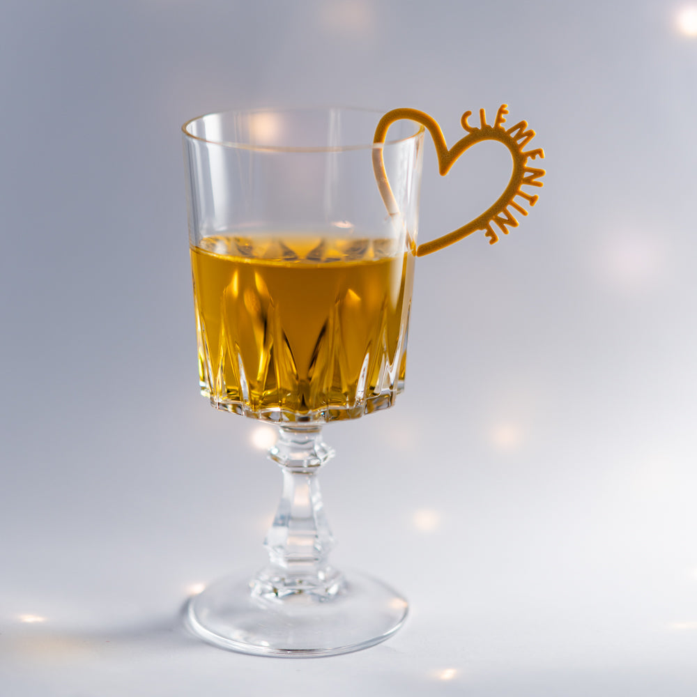 Photo d’un verre à pied en cristal taillé, rempli d’une boisson dorée. Clipé au bord, un marque-verre en forme de cœur couleur doré porte le prénom « Clémentine ». La pièce fine et élégante ajoute une touche romantique et festive à la présentation du verre.