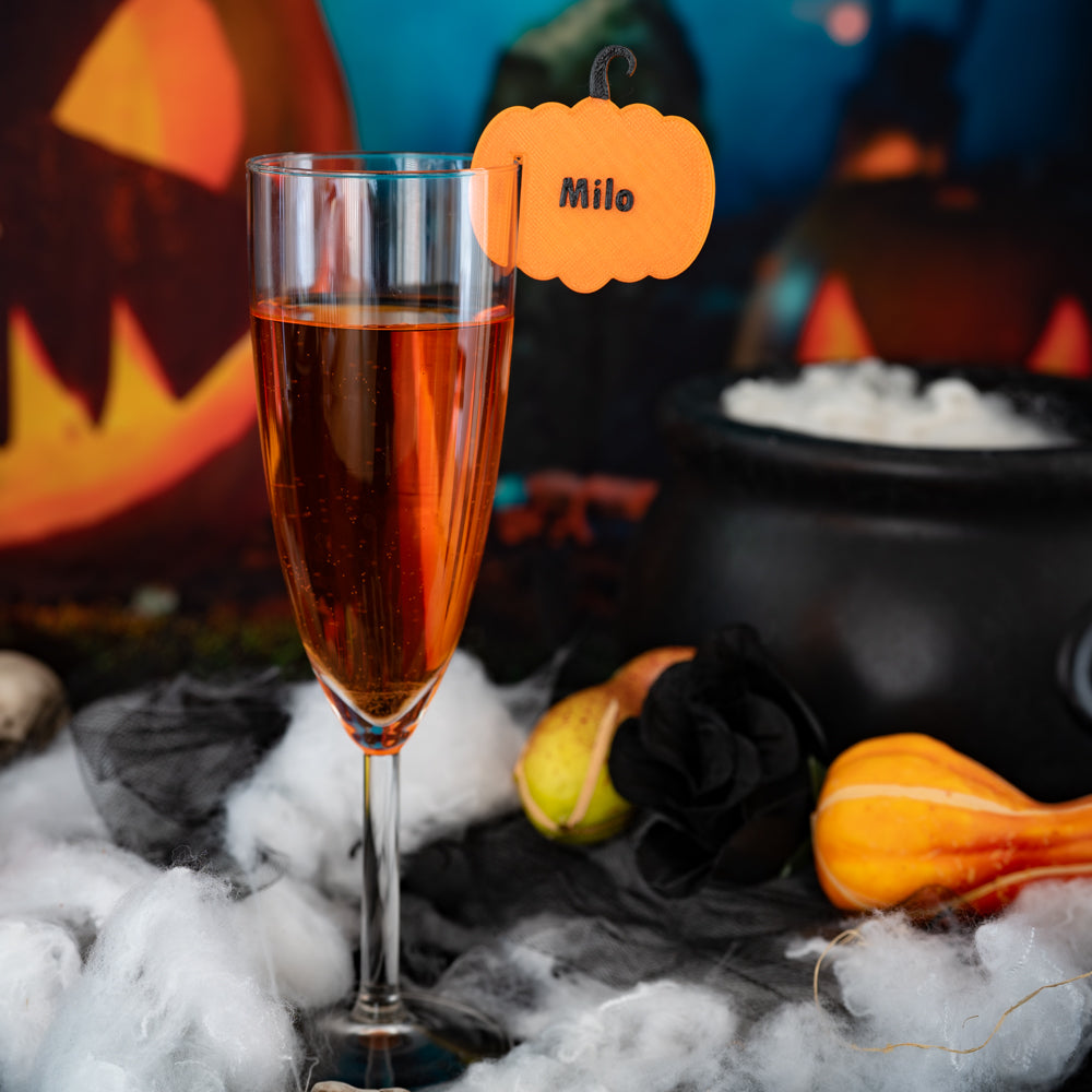 Flûte de cocktail décorée d’un marque-verre orange en forme de citrouille avec le prénom “Milo”, dans une ambiance Halloween.