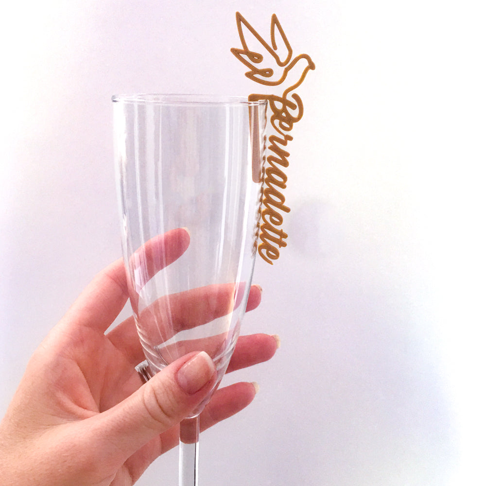 Main tenant une flûte transparente décorée d’un marque-verre doré en forme de colombe avec le prénom “Bernadette”, sur fond clair et lumineux.