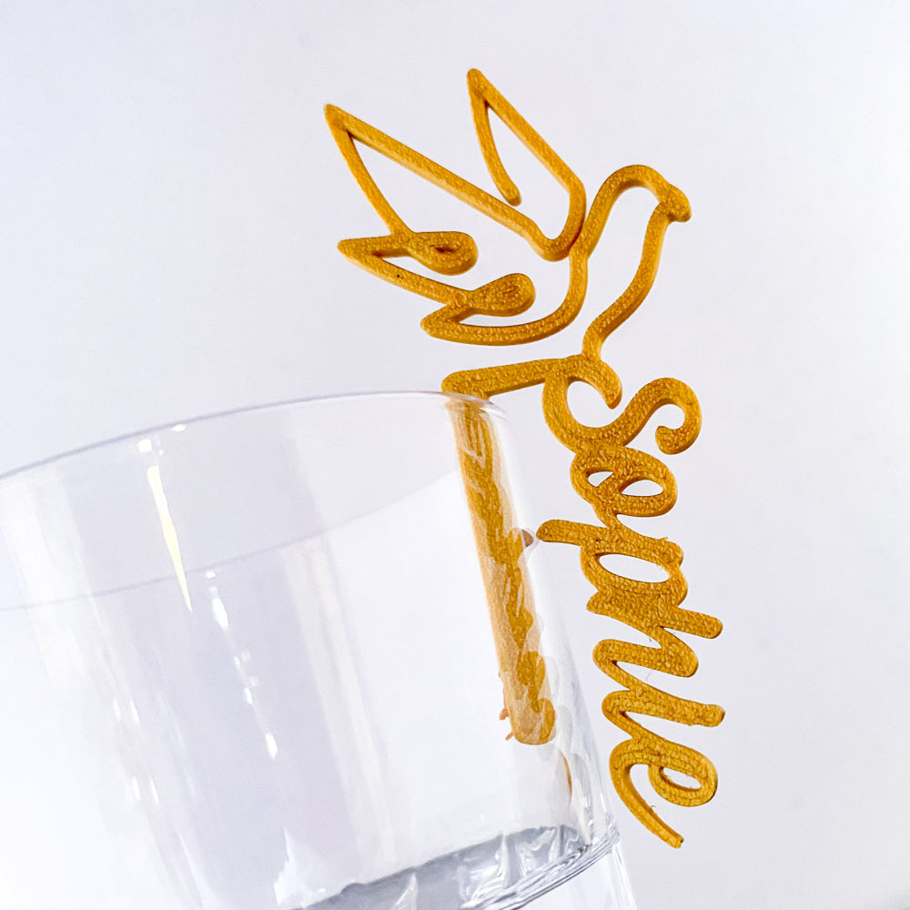 Marque-verre doré en forme de colombe portant le prénom “Sophie”, accroché au bord d’un verre transparent.